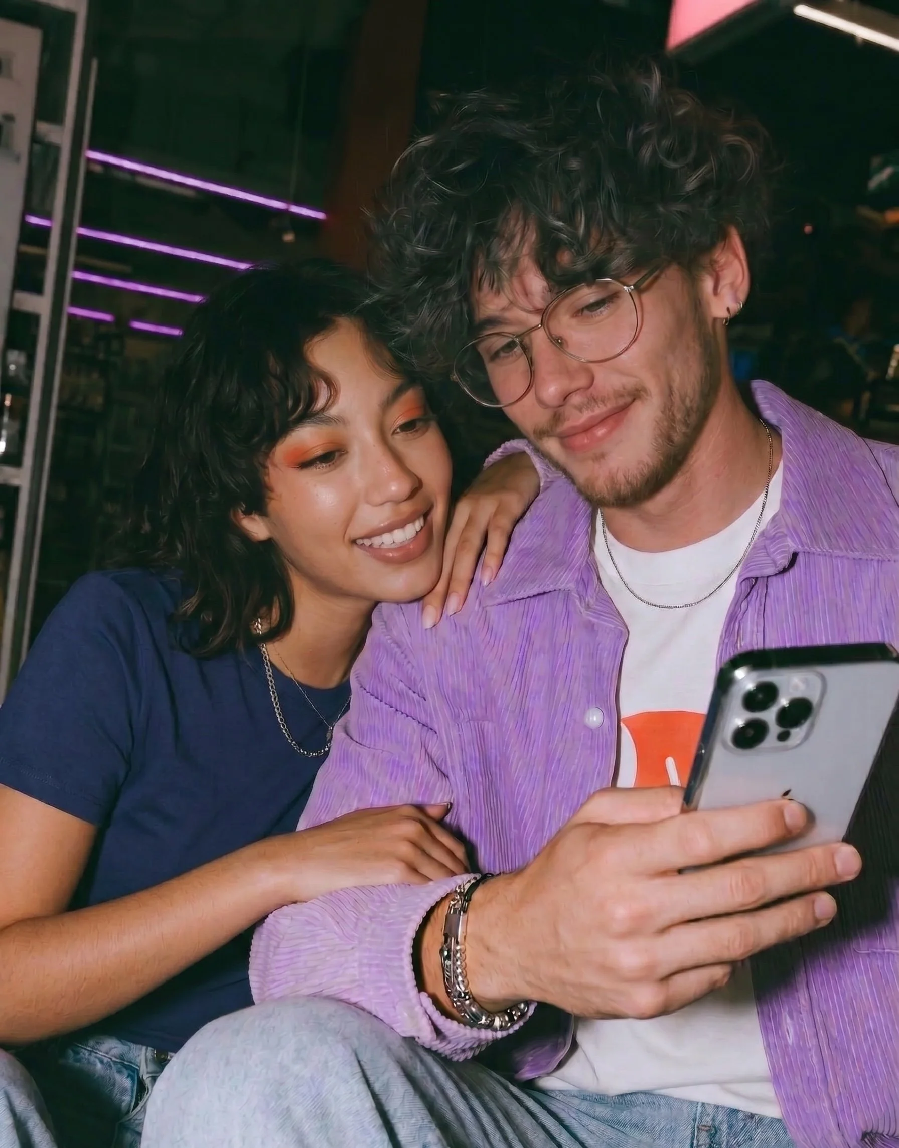 A young woman and a young man taking a selfie together. The woman has dark, curly hair and is smiling, while the man has curly hair, glasses, and is also smiling. They are indoors with neon lighting in the background.