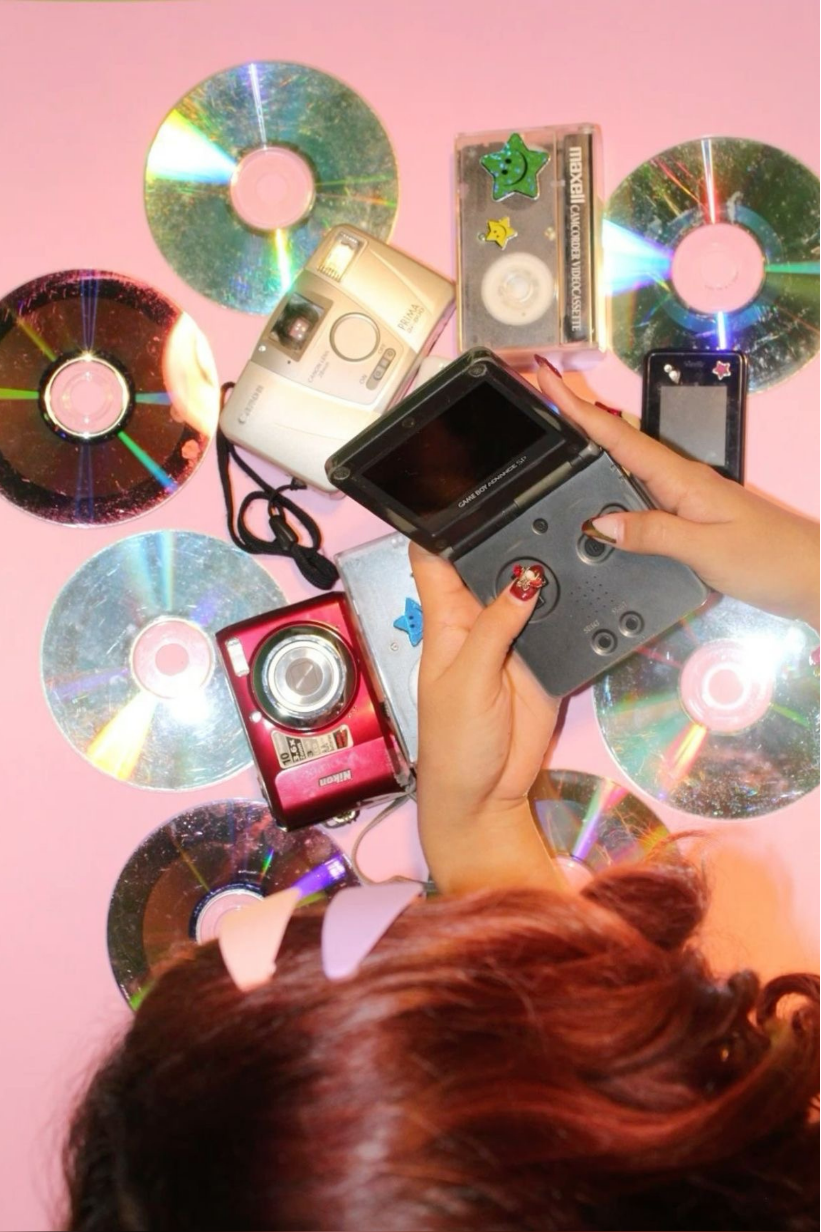 A pink surface with several CDs scattered across it, along with a small vintage camera, a handheld gaming device, and a few stickers. Two hands are visible, one holding the gaming device.