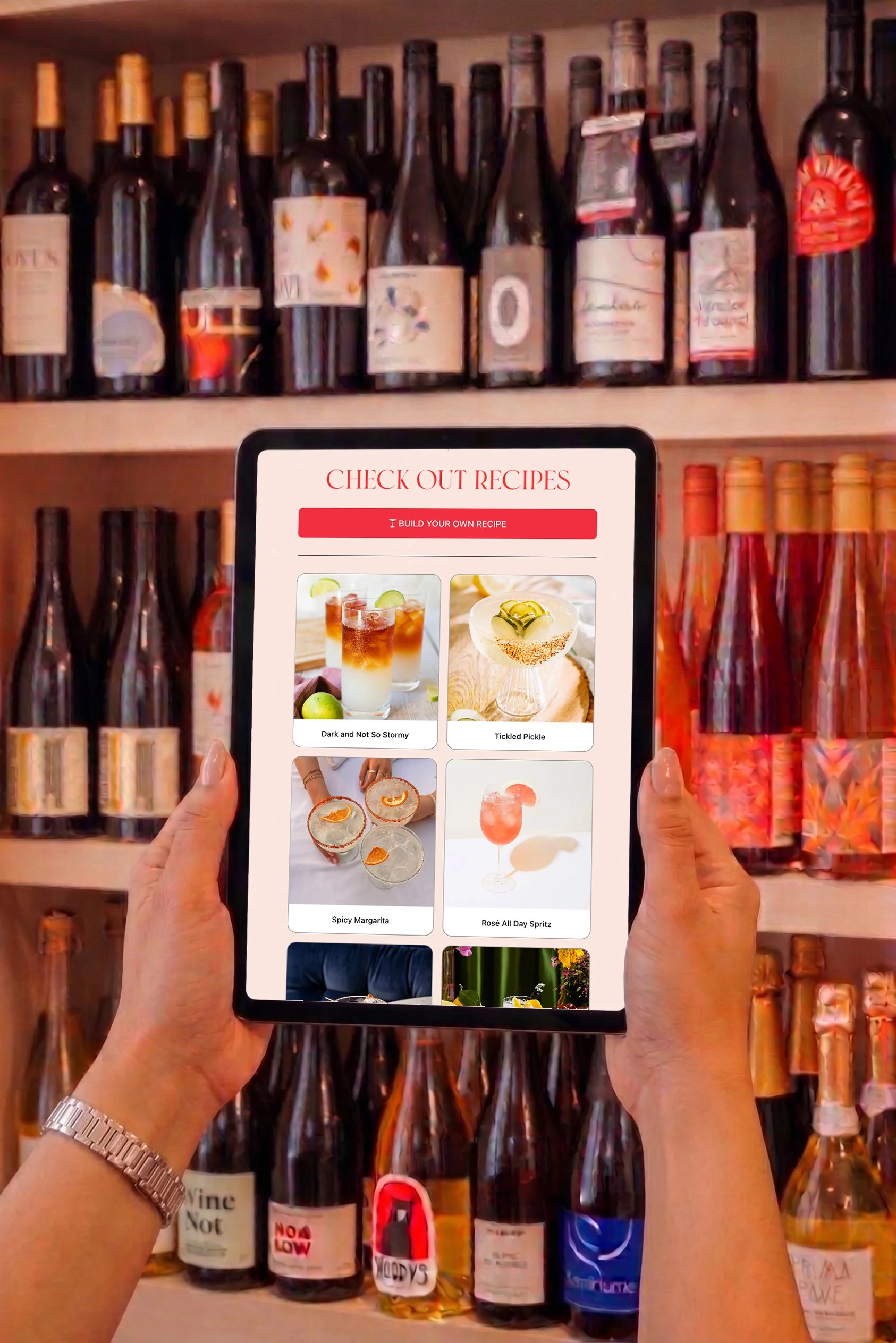 Person holding a tablet displaying cocktail recipes in front of a shelf of wine bottles.