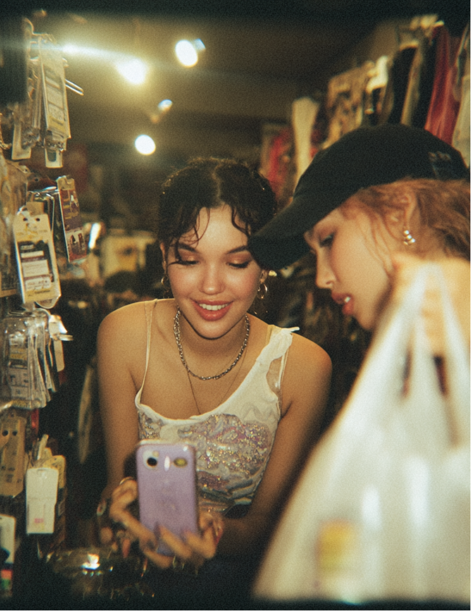 Two young women taking a selfie in a store aisle with bright overhead lights. One woman with short dark hair and a sleeveless top, and another with blonde hair and a black baseball cap. Both appear to be smiling.