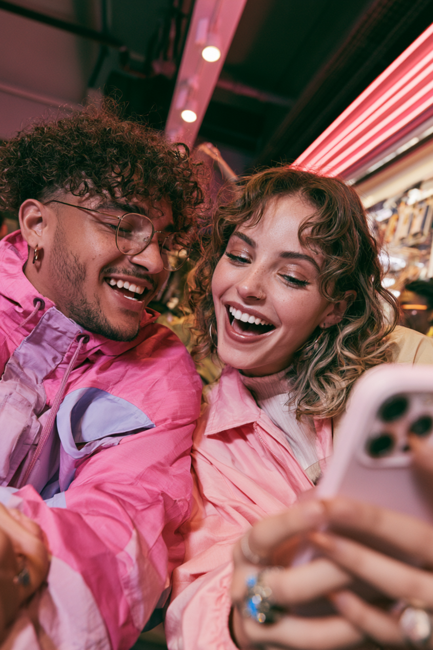 Two young people taking a selfie at a brightly lit outdoor venue. They are smiling, with the man wearing glasses and a pink jacket, and the woman with curly hair and a pink jacket. The background shows a neon-lit setting.