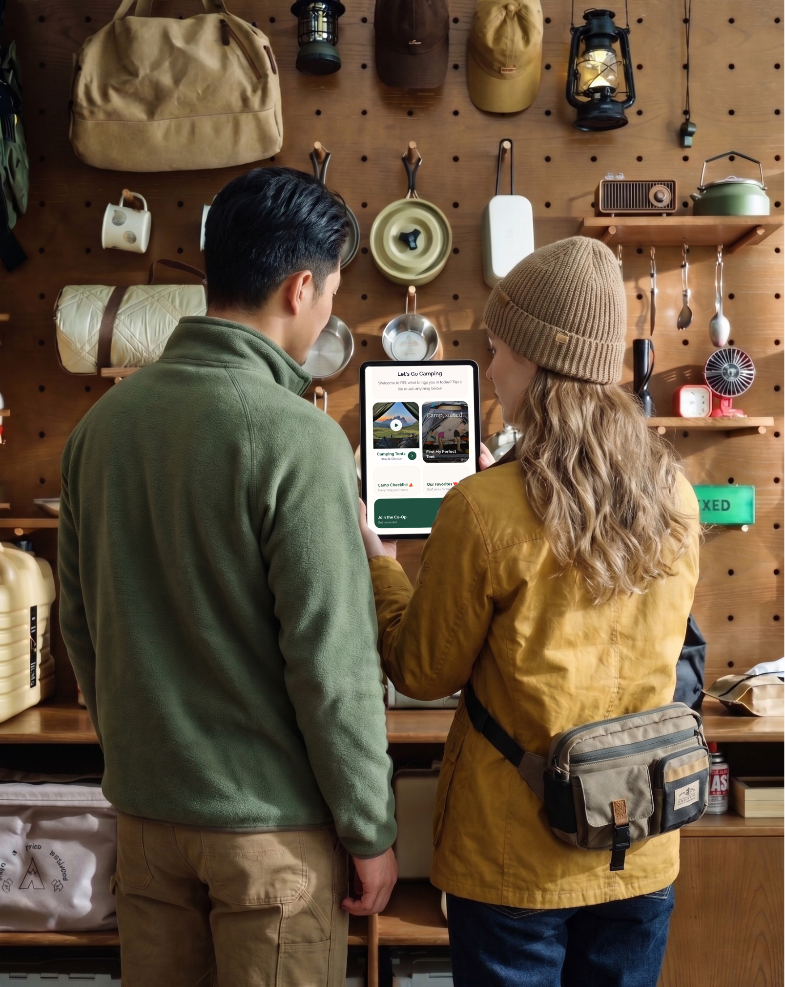 Two people stand in front of a wooden wall with camping gear hanging, including hats, lanterns, cookware, a small shelf, and utensils, as they look at a tablet.