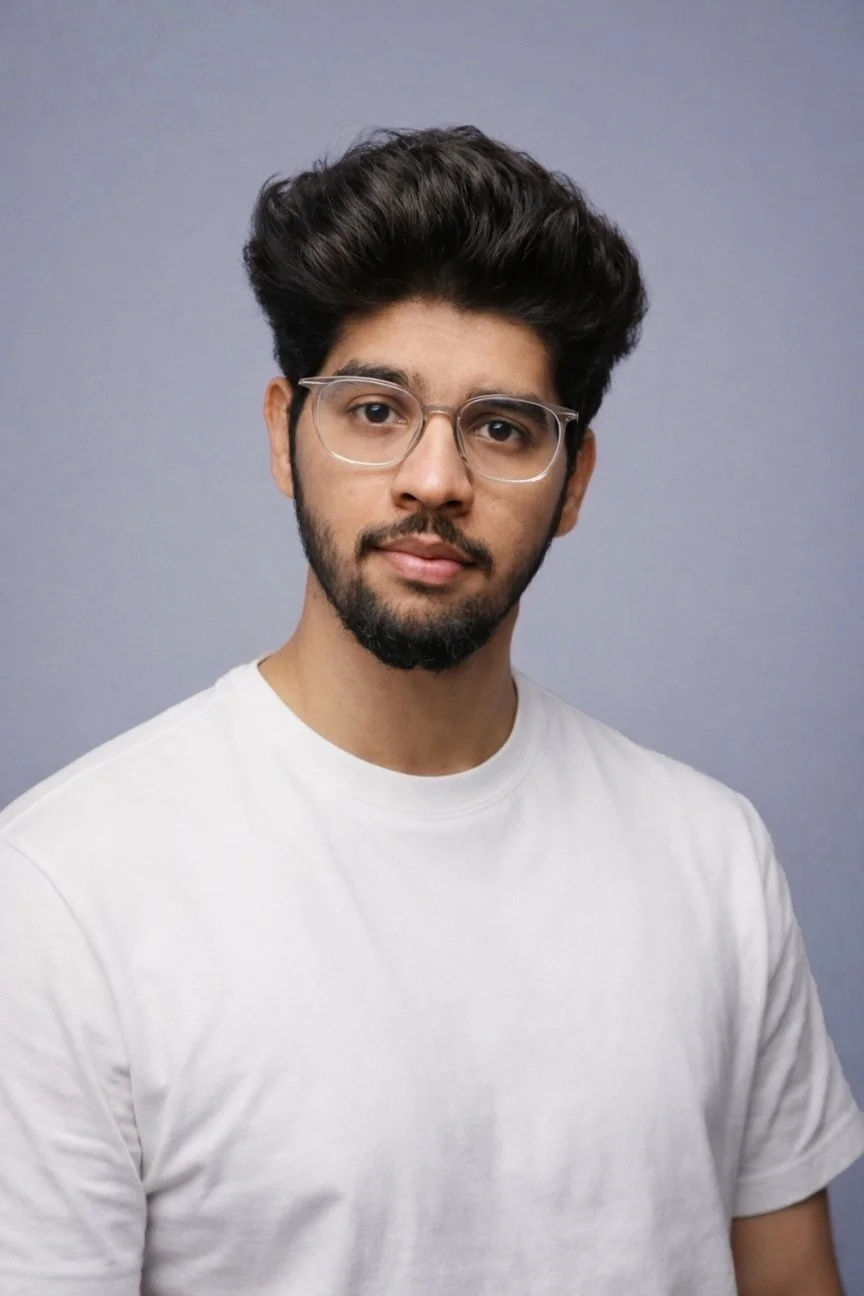 A young man with dark hair, beard, and glasses wearing a white t-shirt, standing against a plain gray background.