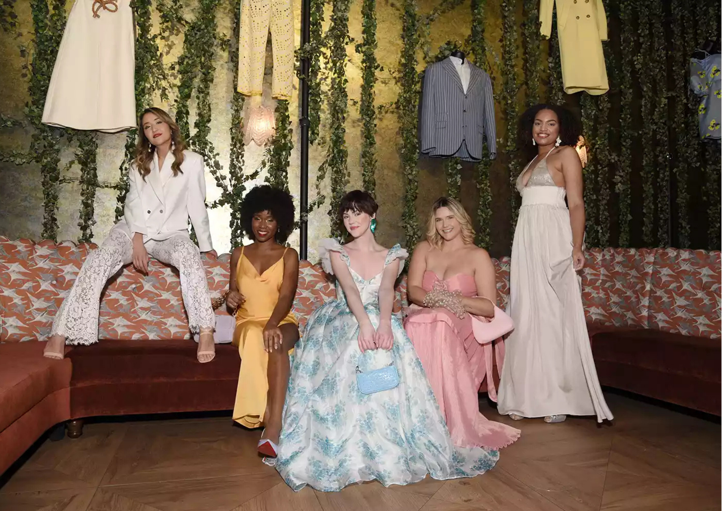 Six women posing together in a room with hanging clothes and green ivy decor, sitting and standing on a patterned orange and white sofa.
