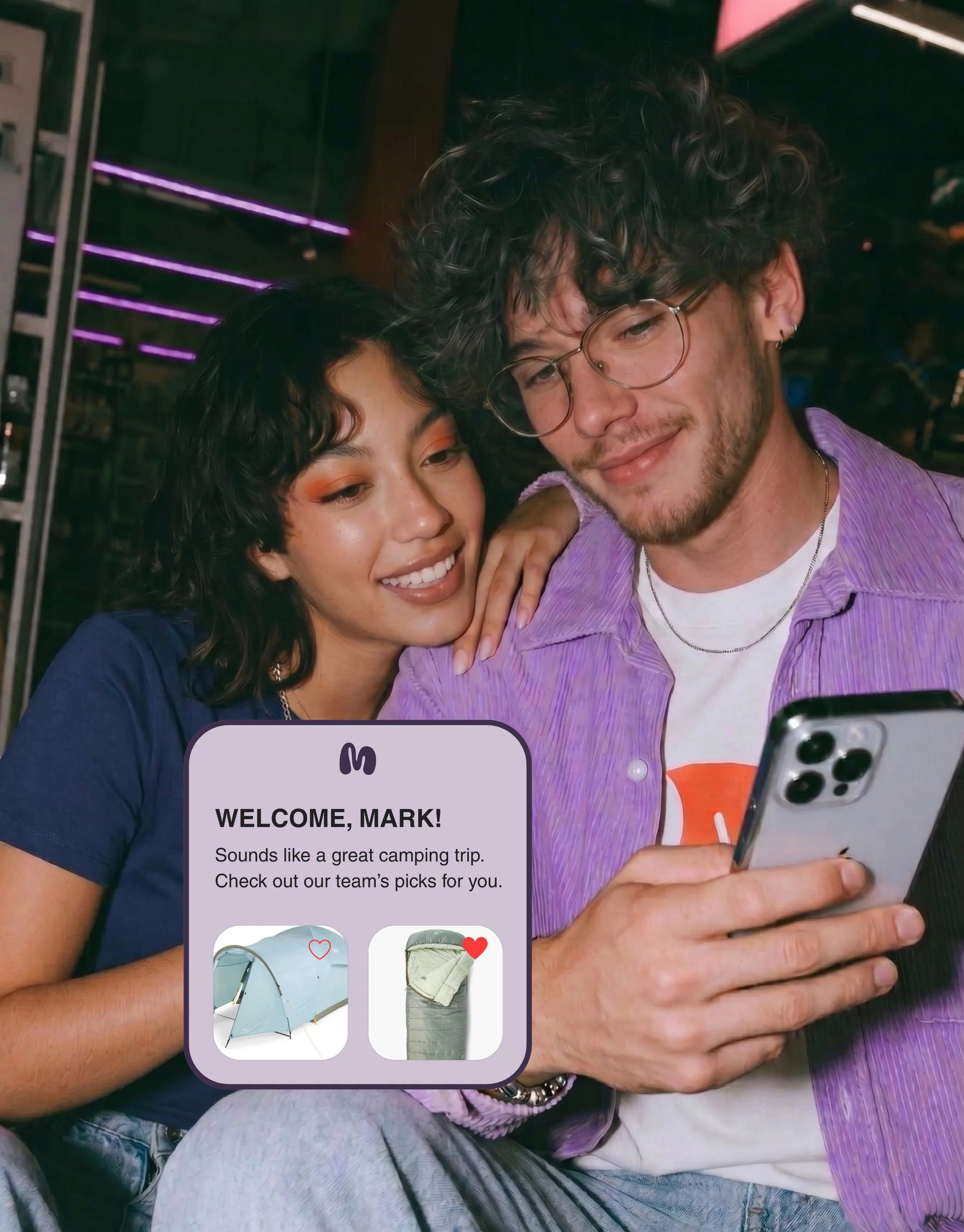 Two friends, a woman with dark curly hair and a man with light curly hair and glasses, are smiling and taking a selfie. The woman is wearing a navy blue shirt, and the man is wearing a purple shirt and a white t-shirt underneath. They are in a cozy indoor space with purple neon lights in the background.