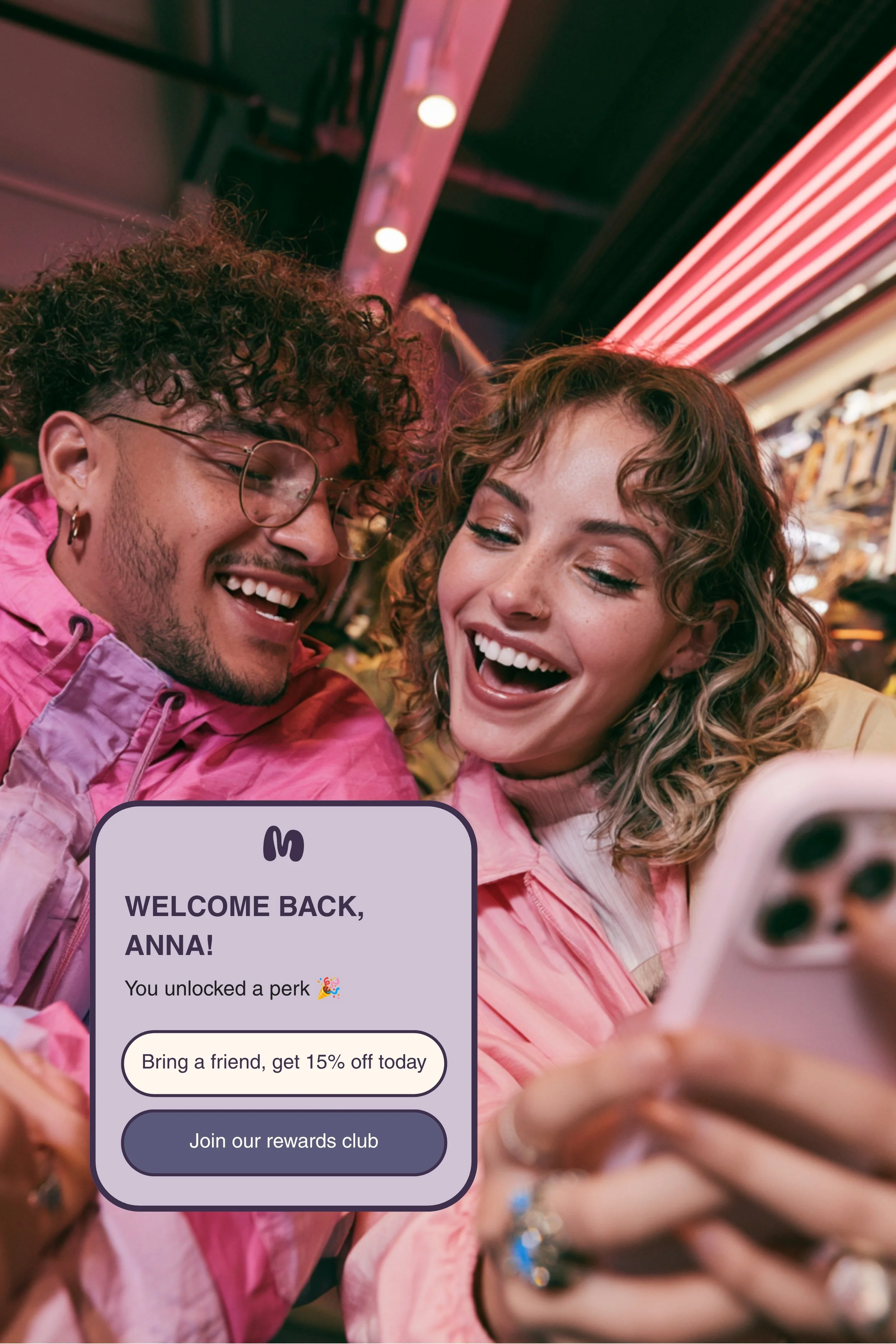 Two young adults, a man and a woman, smiling and taking a selfie at a vibrant, likely indoor location with colorful lighting and a striped awning above them. They are dressed in pink, and the woman is holding a smartphone. A text overlay in the image reads, "Welcome back, Anna! You unlocked a perk 🎉. Bring a friend, get 15% off today. Join our rewards club."