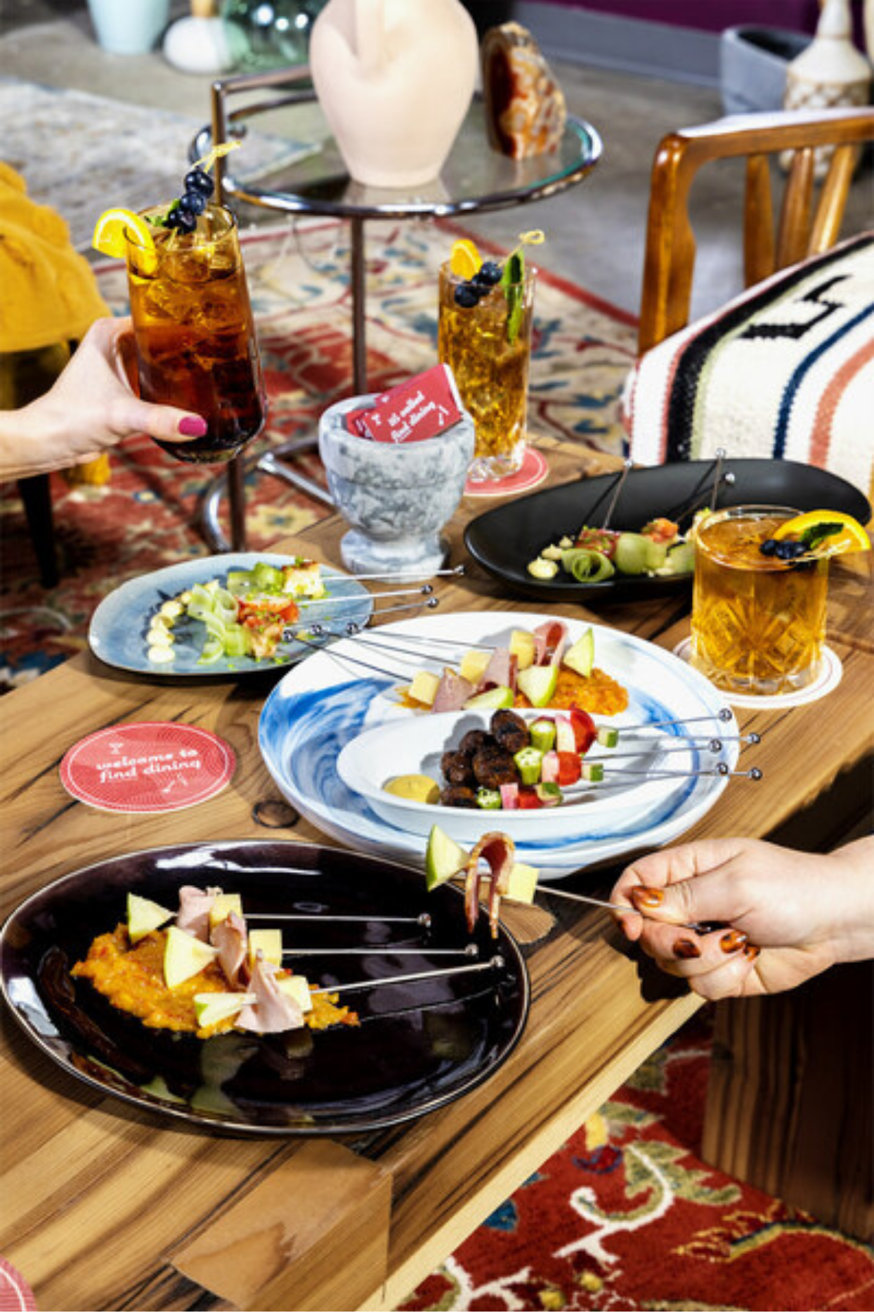 People enjoying food and drinks at a social gathering, with various appetizers, skewers, and cocktails on a wooden table.