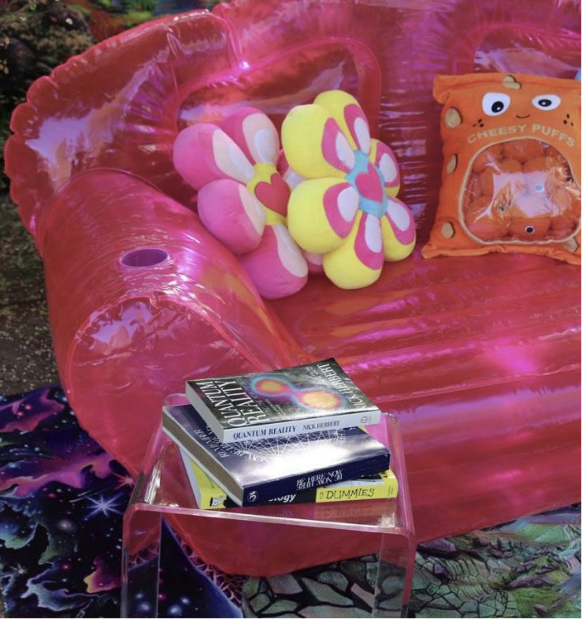 A pink inflatable pool with pink and yellow water balloons, a box of 'Quantum Reality' card game, and a bag of cheese puffs in a colorful setting.