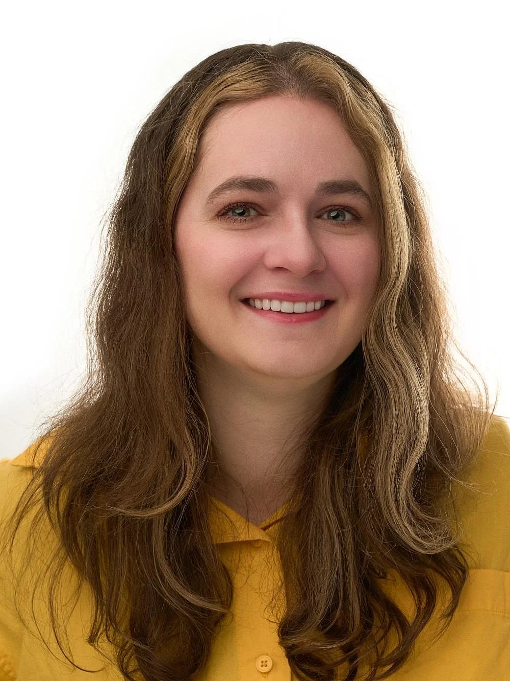 A young woman with long wavy brown hair smiling, wearing a yellow top, against a plain white background.
