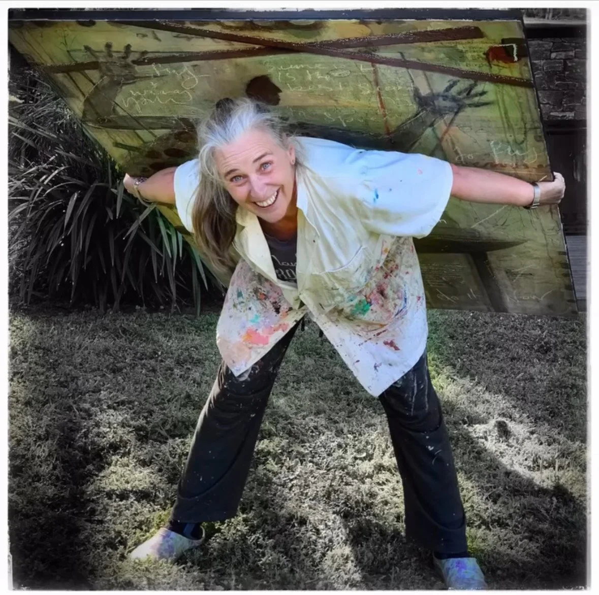 A woman with long gray hair, smiling, leaning forward with arms outstretched holding a large, painted wooden board behind her. She is wearing a white paint-splattered shirt and black pants, standing outdoors on a grassy area. Her shirt and pants are also covered in paint splatters.