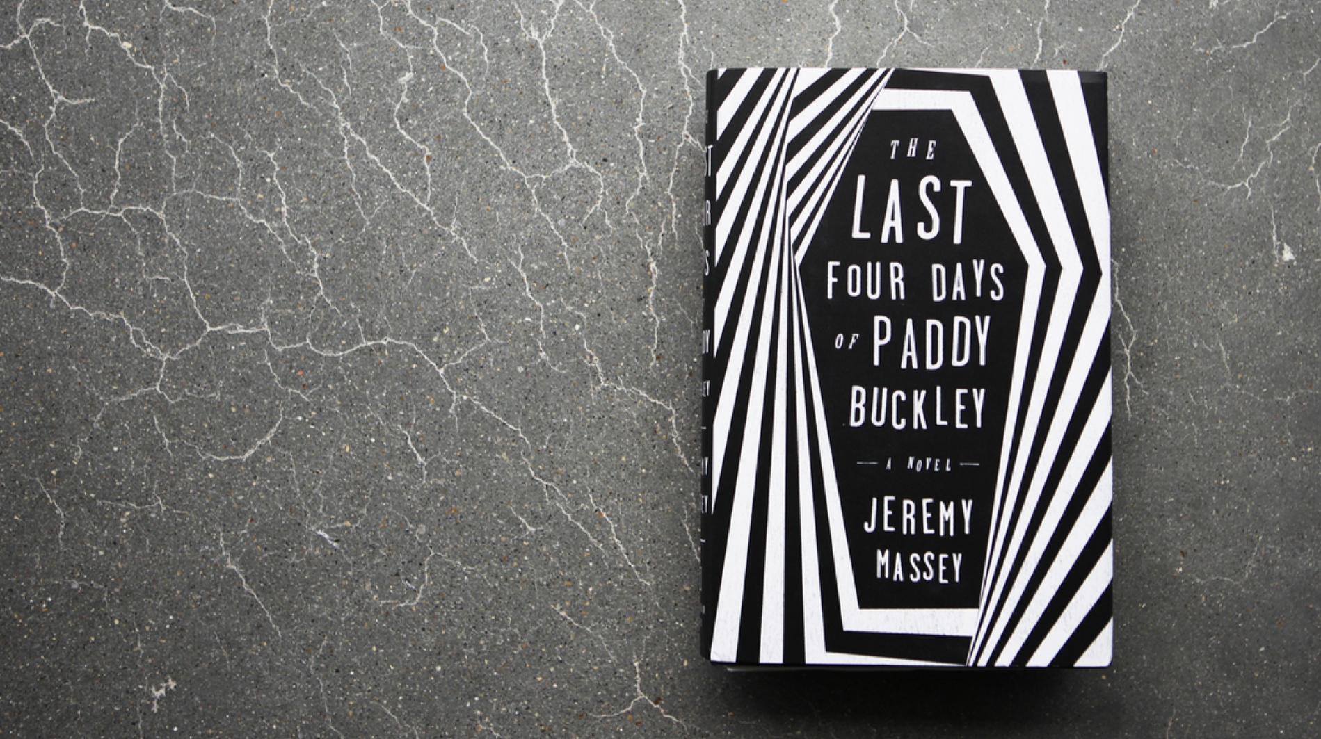 The image shows a black and white patterned book cover titled "The Last Four Days of Paddy Buckley" by Jeremy Massey, lying on a cracked concrete surface.