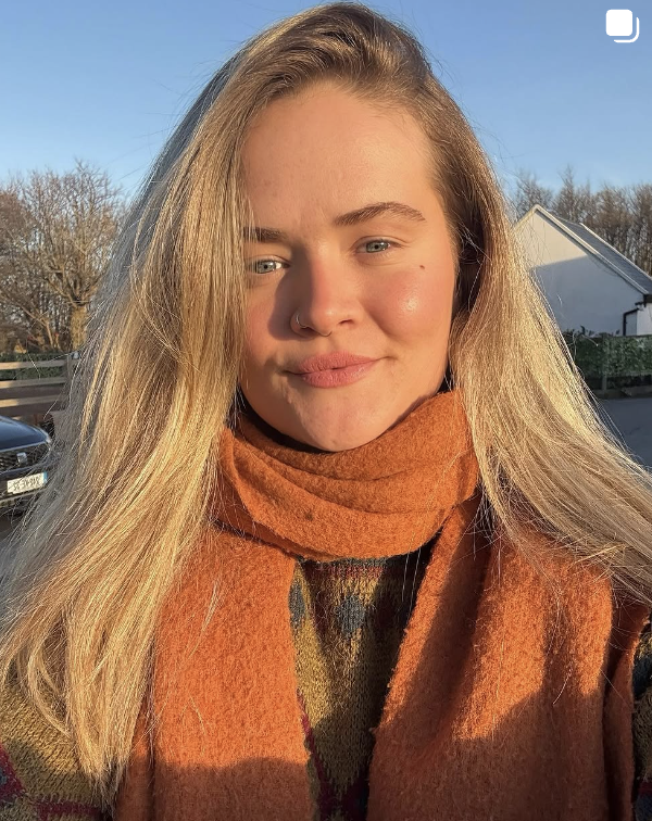 A young woman with long blonde hair and a nose ring, smiling outdoors on a sunny day, wearing an orange scarf and a sweater.