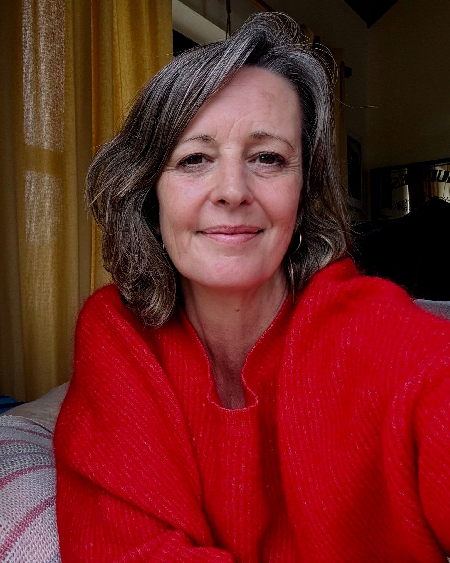 Close-up of a middle-aged woman smiling, wearing a red sweater, with brown and gray hair, sitting indoors near yellow curtains.