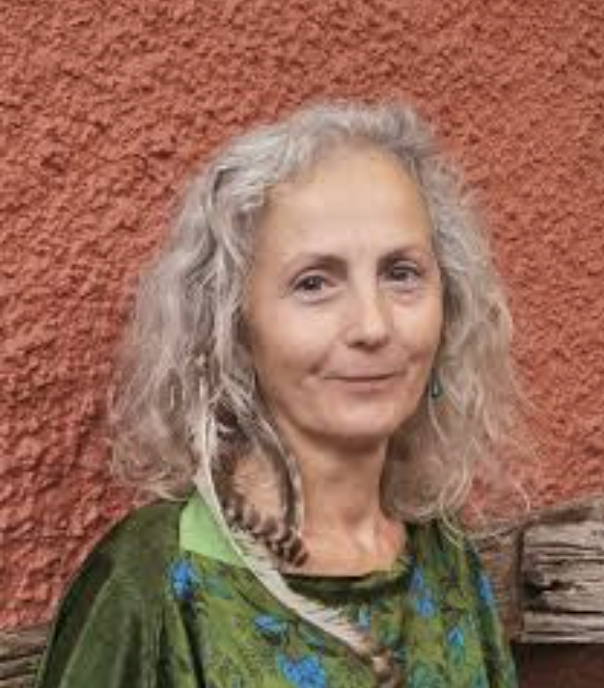 A woman with curly gray hair standing in front of a textured red wall, wearing a green top with a floral pattern.