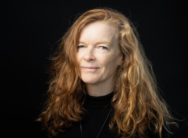 Portrait of a woman with long, curly red hair, wearing a black top, set against a dark background.