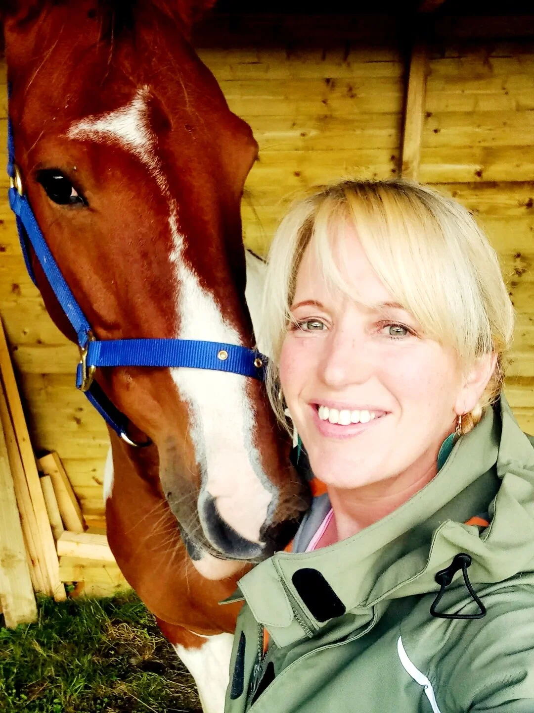 A woman with blonde hair taking a selfie with a brown horse with white markings on its face inside a wooden stable.
