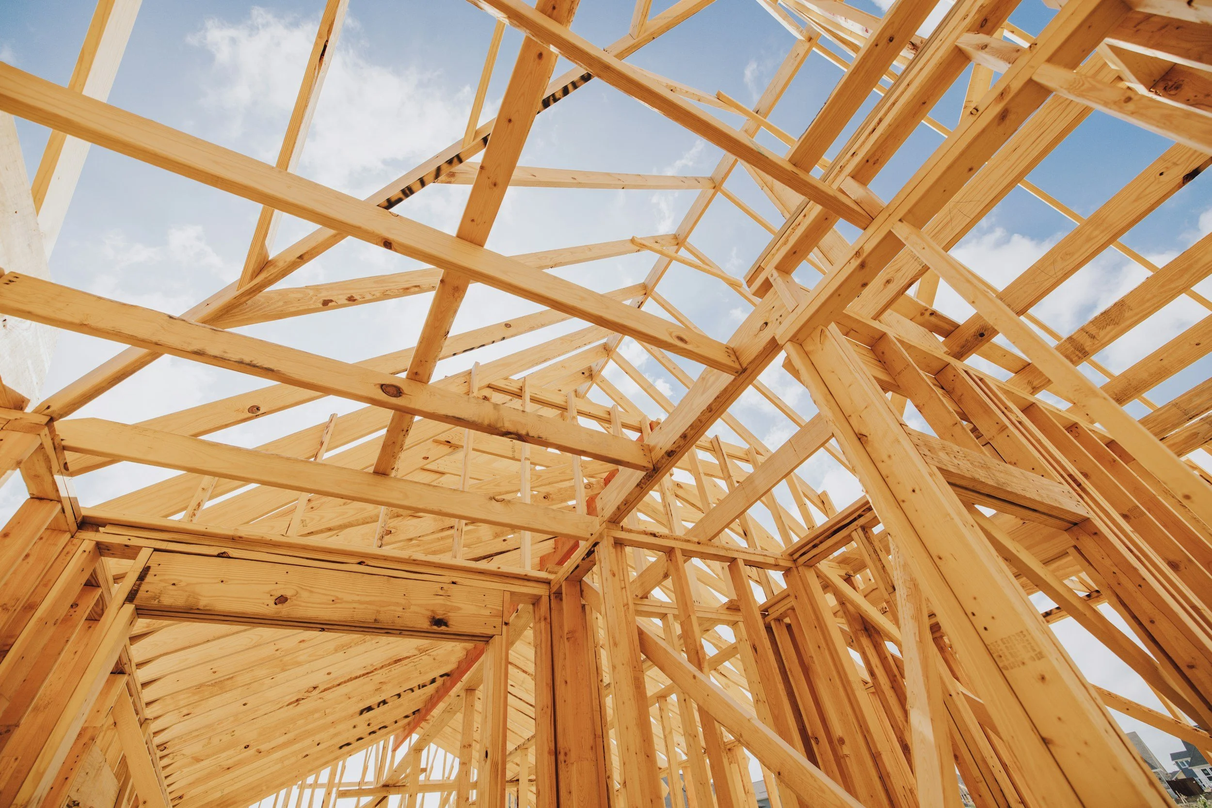 Wooden framing structure of a building under construction, viewed from inside with blue sky and clouds in the background.