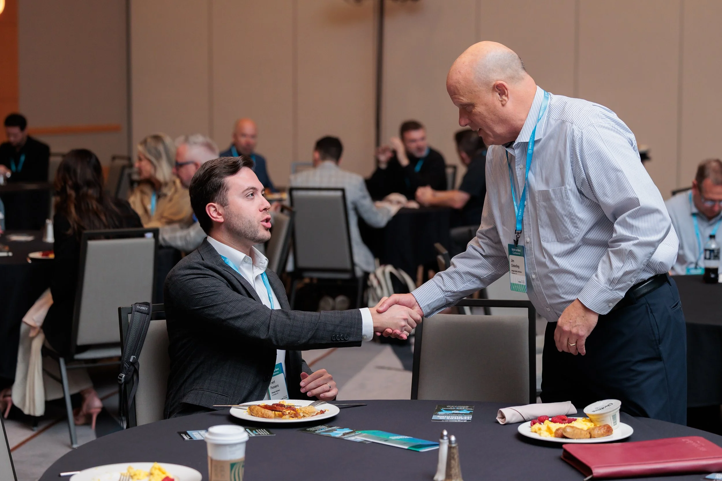 Whelan Advisory team sitting at a conference table shaking hands with an older man who is standing. In the background, there are several other people engaged in conversations. The table has plates of food, coffee cups, and conference materials.