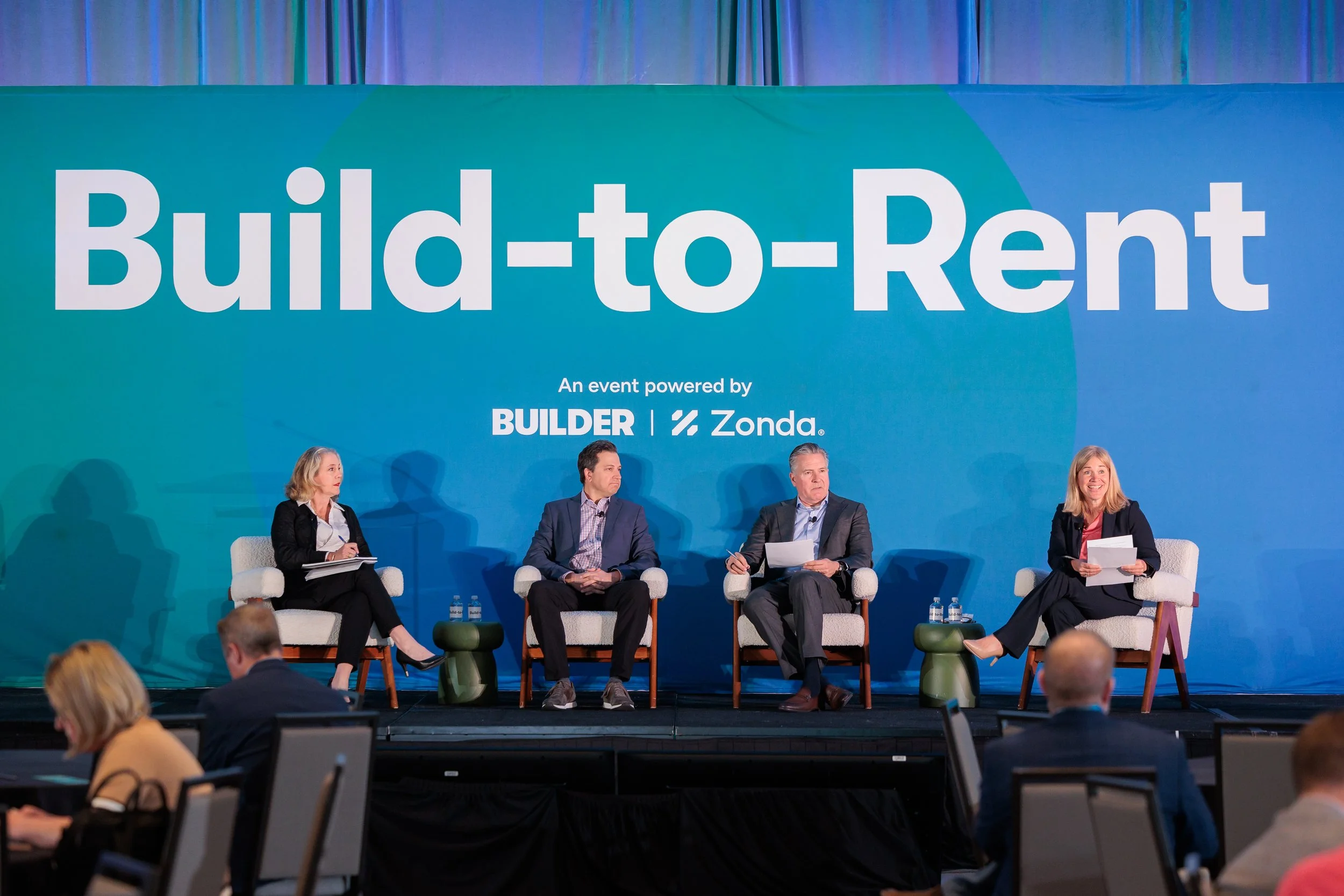 A panel of four people seated on stage during a conference or panel discussion, with a blue backdrop displaying the words 'Build-to-Rent' and logos of BUILDER and Zonda. Audience members are visible in the foreground.