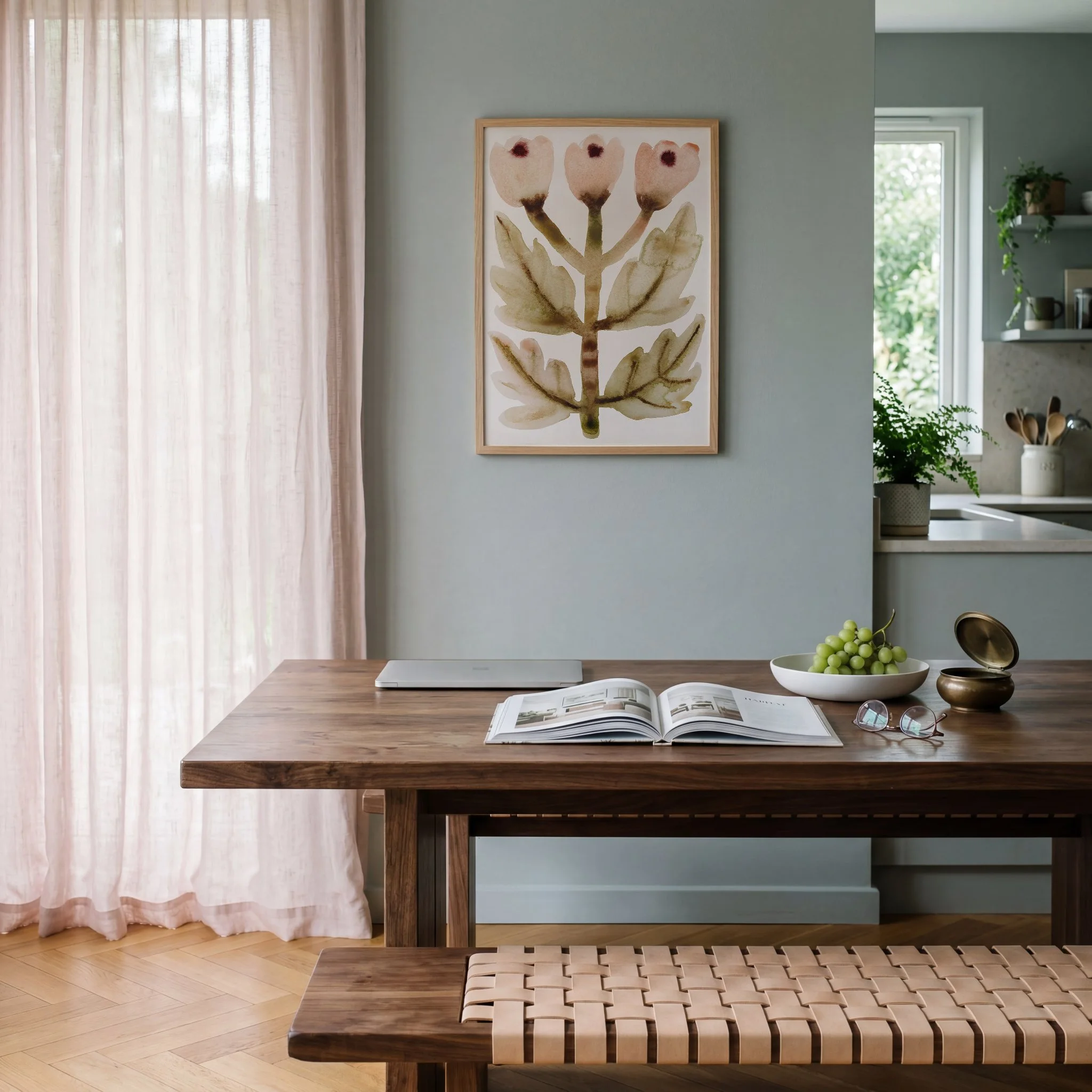 A dining table with a light wood woven bench, an open magazine, a pair of glasses, and a white bowl of green grapes. In the background are pale pink curtains, a framed botanical watercolor artwork of a plant with three pink flowers, a window with greenery outside, and a shelf with potted plants and other decor.
