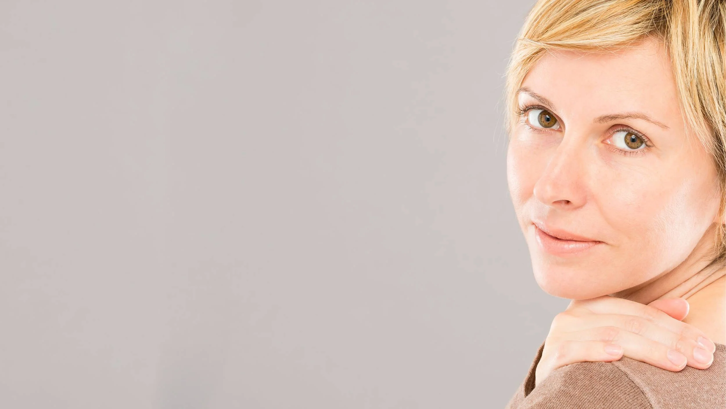 Close-up of a woman with blonde hair and light skin resting her chin on her hand, against a plain gray background.