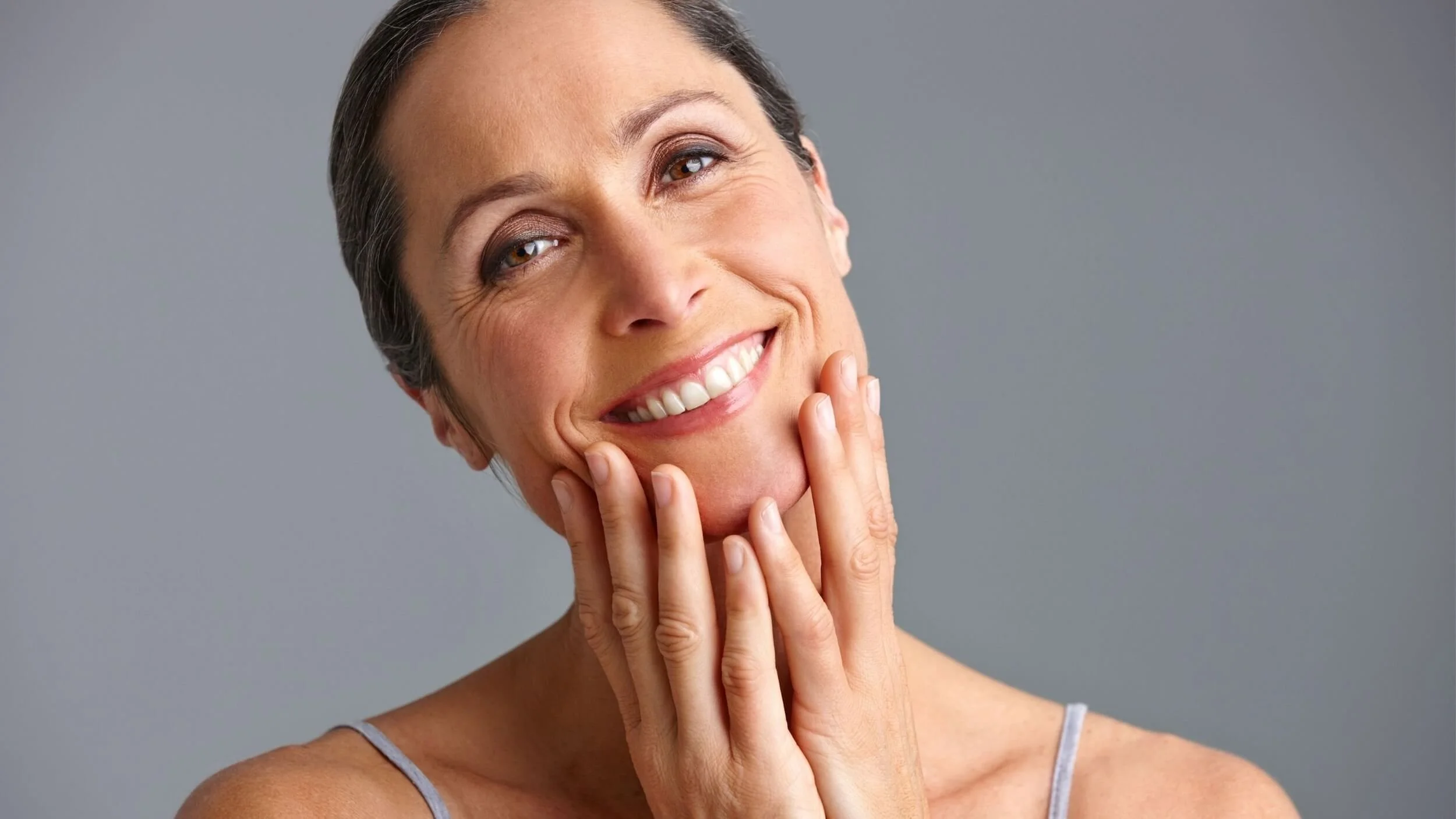 A smiling middle-aged woman with short gray hair touching her face with her hands, wearing a sleeveless top, against a plain gray background.