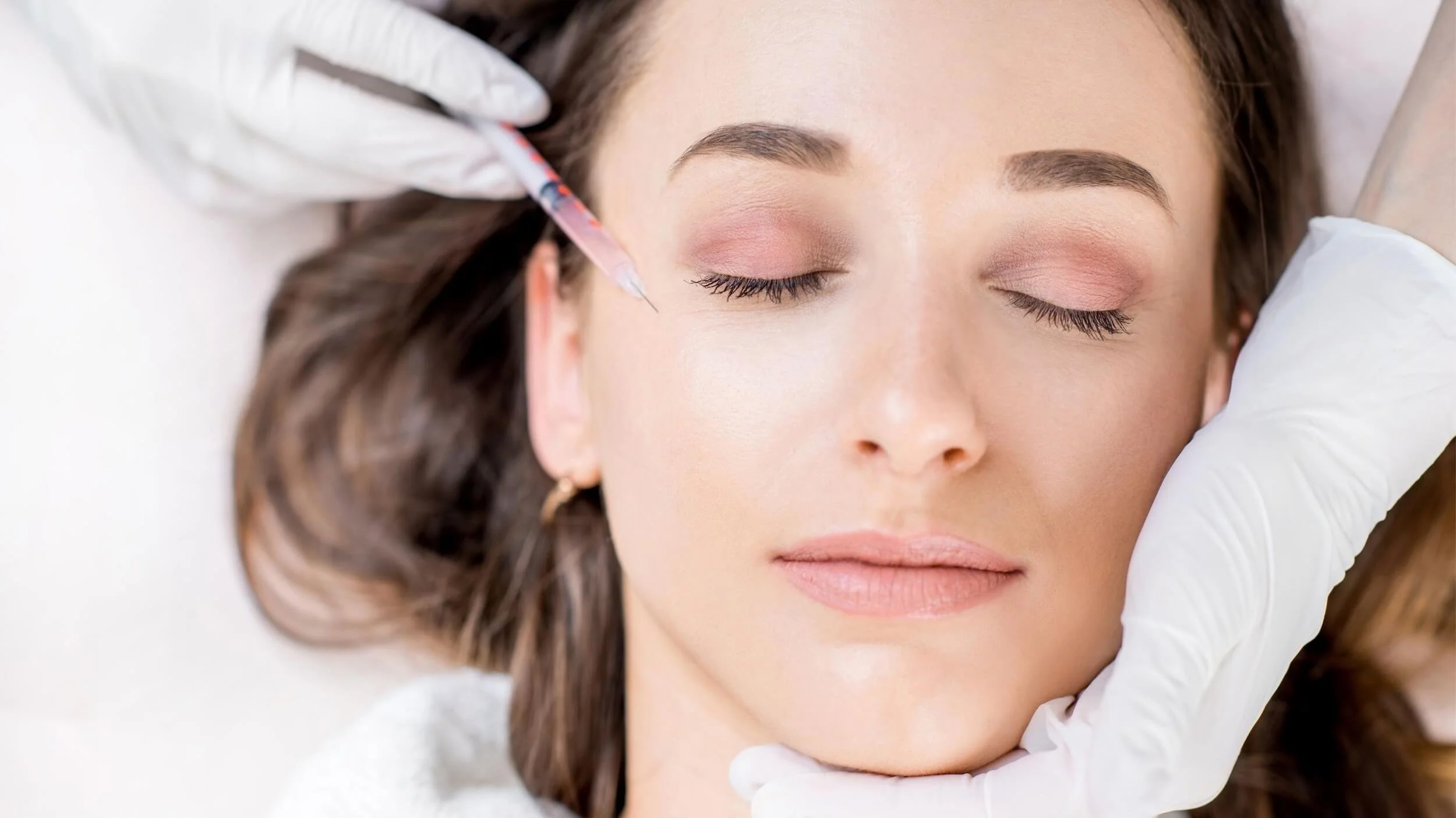 A woman with closed eyes receiving a cosmetic injection near her eyebrow, with a person wearing latex gloves administering the treatment.