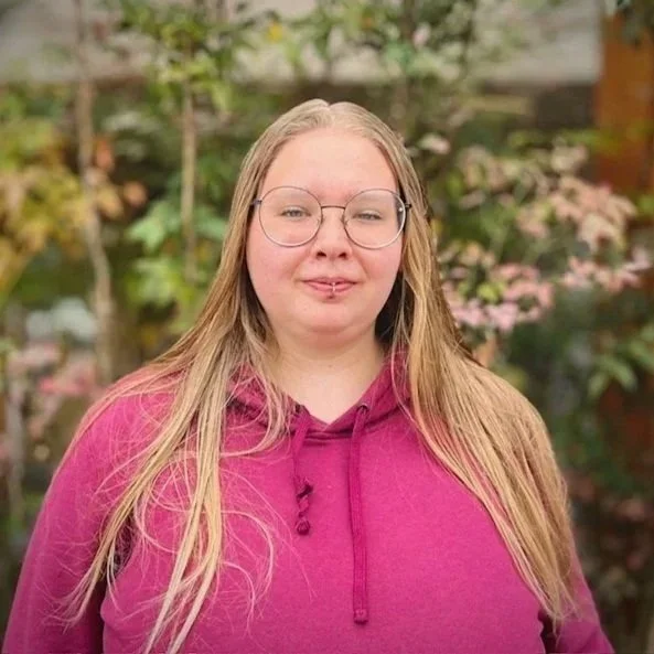 Young woman with light skin, red glasses, and blonde hair tied back, standing outdoors in front of a leafy background, wearing a black shirt and a light plaid shirt.