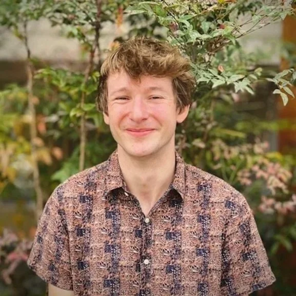 Young man with light brown hair, red glasses, wearing a blue and white striped shirt, smiling outdoors in front of green foliage.
