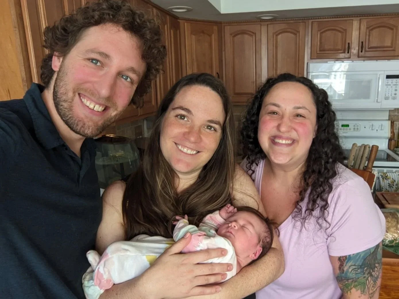 Four people, three women and one man, smiling for a selfie inside a house, holding a newborn baby. A birthday balloon is visible in the background.