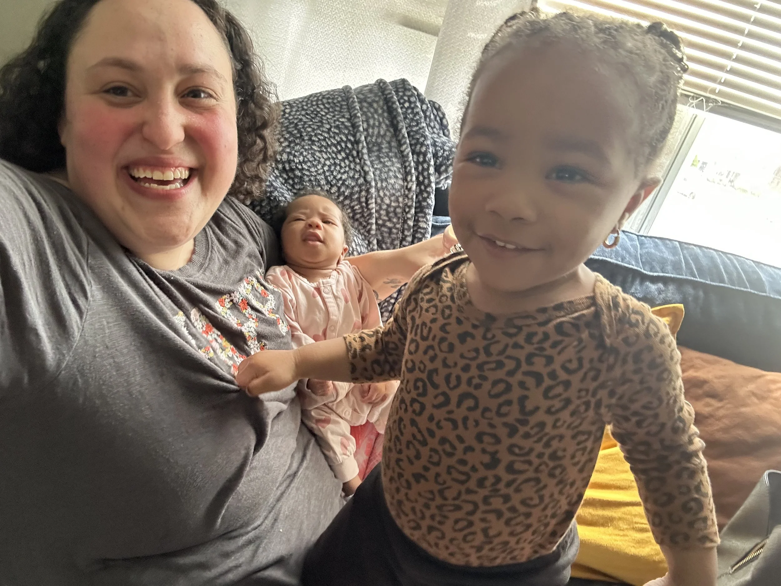 A woman is smiling while holding a baby girl, with a young girl with braided hair and earrings standing in front of them, all smiling in a living room with a couch and window in the background.