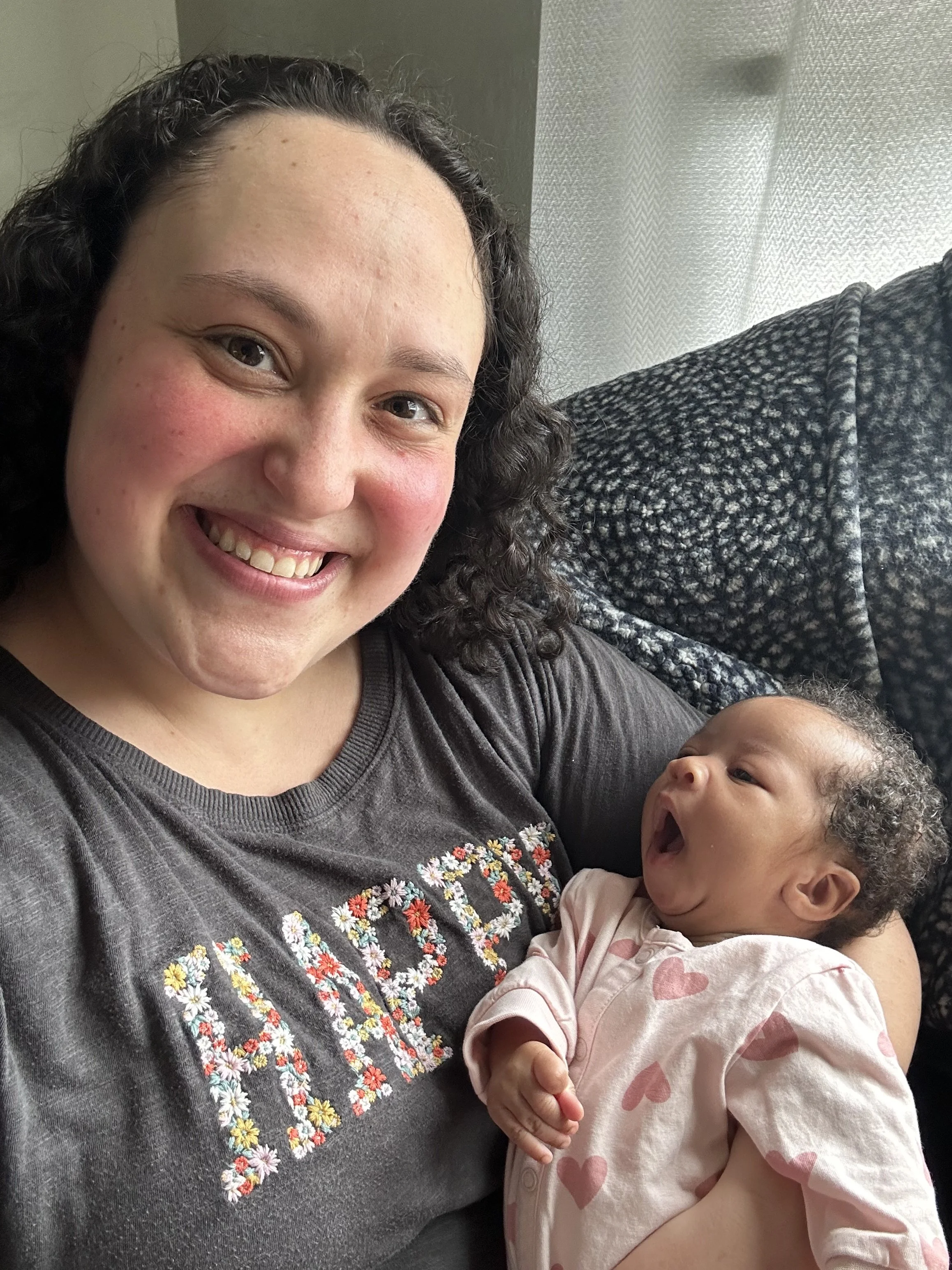 A woman with curly dark hair smiling and holding a baby with curly hair, wearing a pink outfit with heart designs, in a room with a textured gray sofa and light-colored curtains.