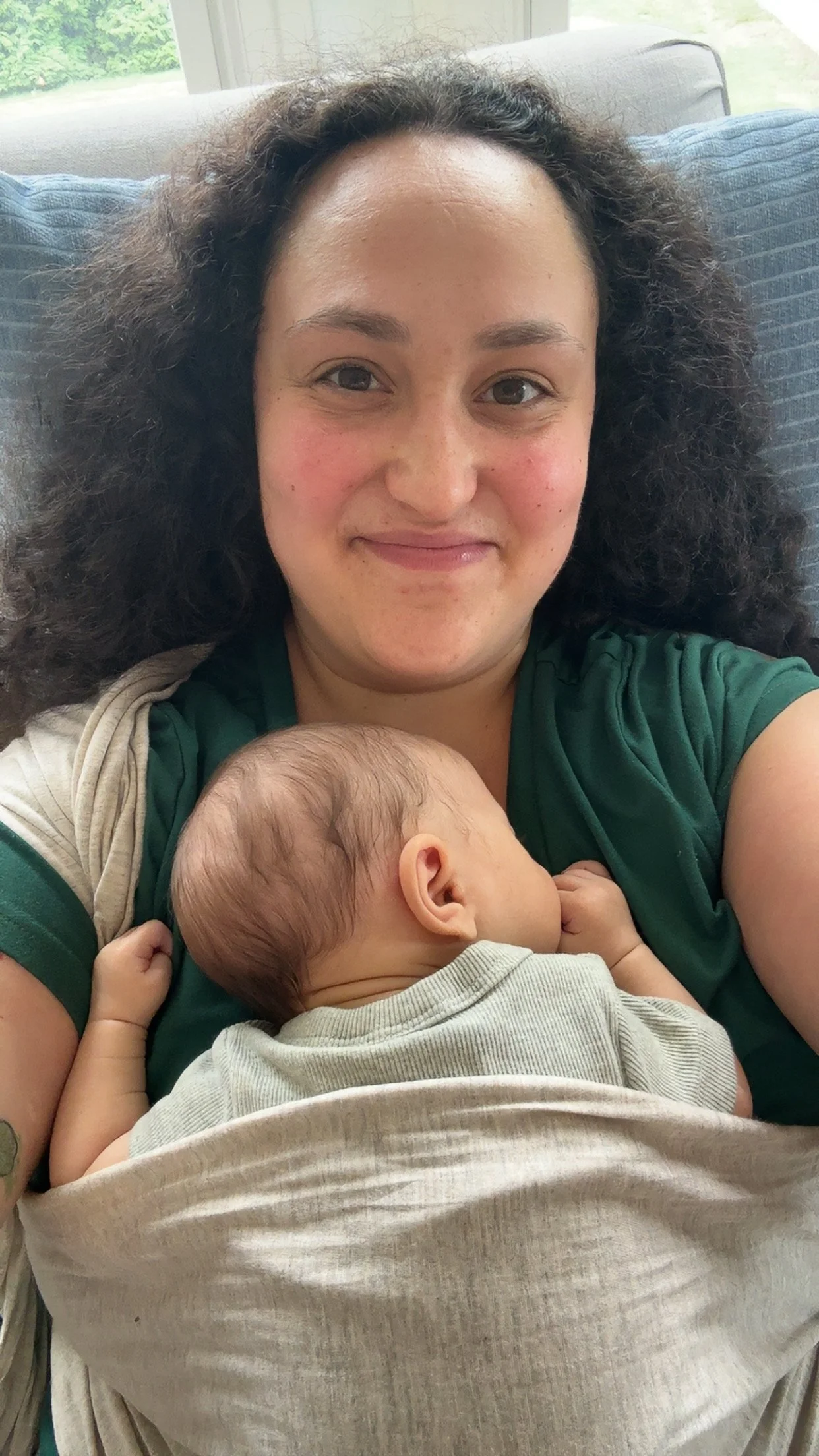 A woman with curly hair smiling while holding a sleeping baby close to her chest.