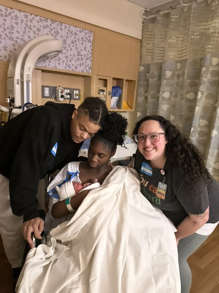 A woman with glasses and curly hair sitting on the bed, smiling, as she holds someone's foot. The bed is messy with blankets, and there is a TV and shelves in the background.
