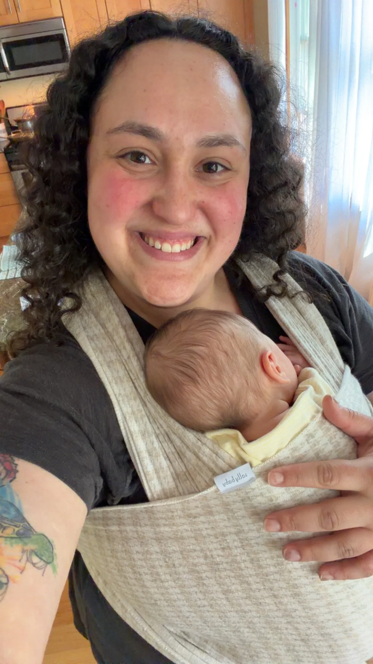 A woman with curly hair and tattoos on her arm is smiling and holding a sleeping baby in a baby carrier. The woman is in a kitchen with wooden cabinets and stainless steel appliances, and there is a window with curtains in the background.