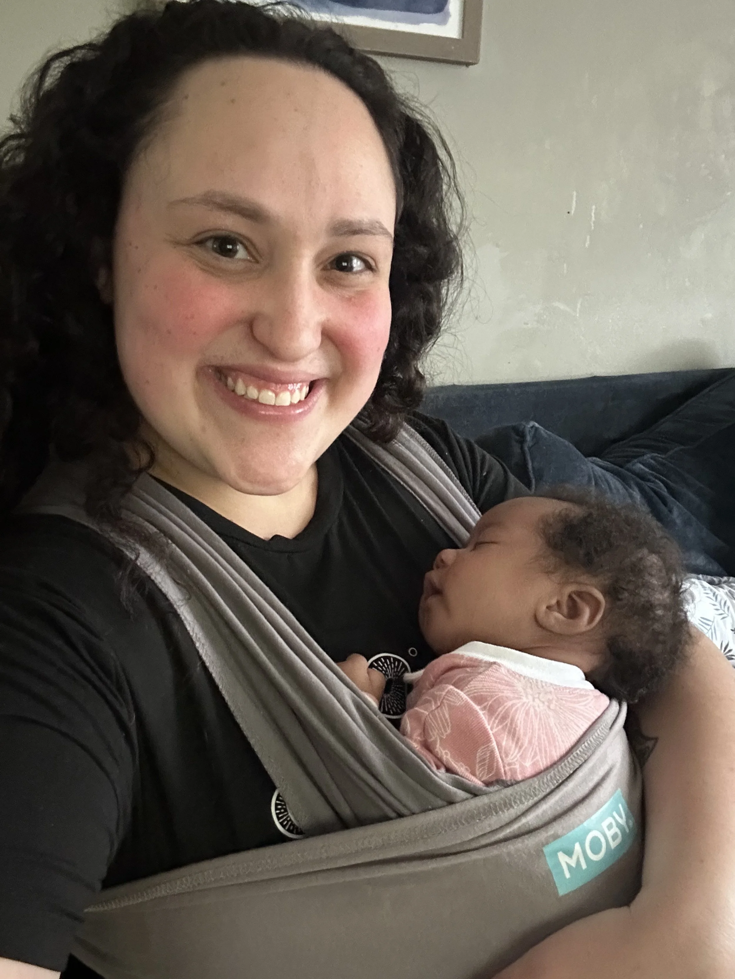 Smiling woman with curly dark hair holding a sleeping baby in a front carrier. The woman is indoors, sitting on a couch with a plain wall behind her.