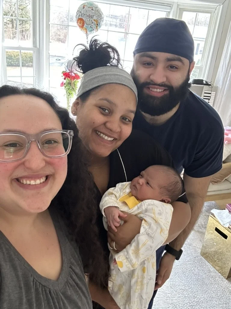 A group of four people, including a woman with glasses, a woman with dark hair tied up, a man wearing a dark head covering, and a newborn baby, all smiling for a selfie inside a bright room with large windows and a birthday balloon in the background.
