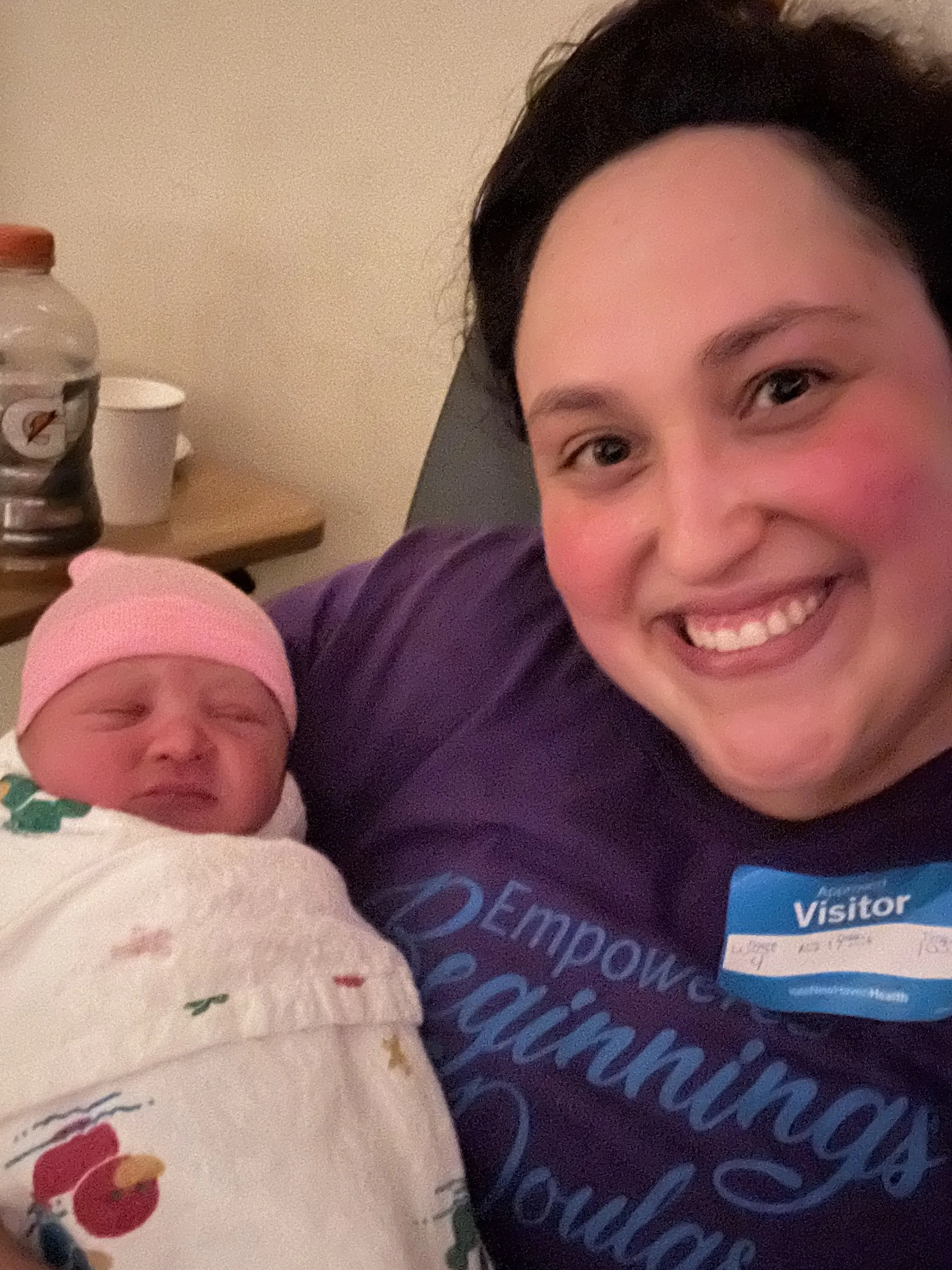 A woman smiling and holding a crying newborn baby, both are close to the camera, with the woman wearing a purple shirt and the baby wrapped in a blanket with a cartoon design, in a room with a beige wall and a table with a gatorade bottle and paper cups in the background.