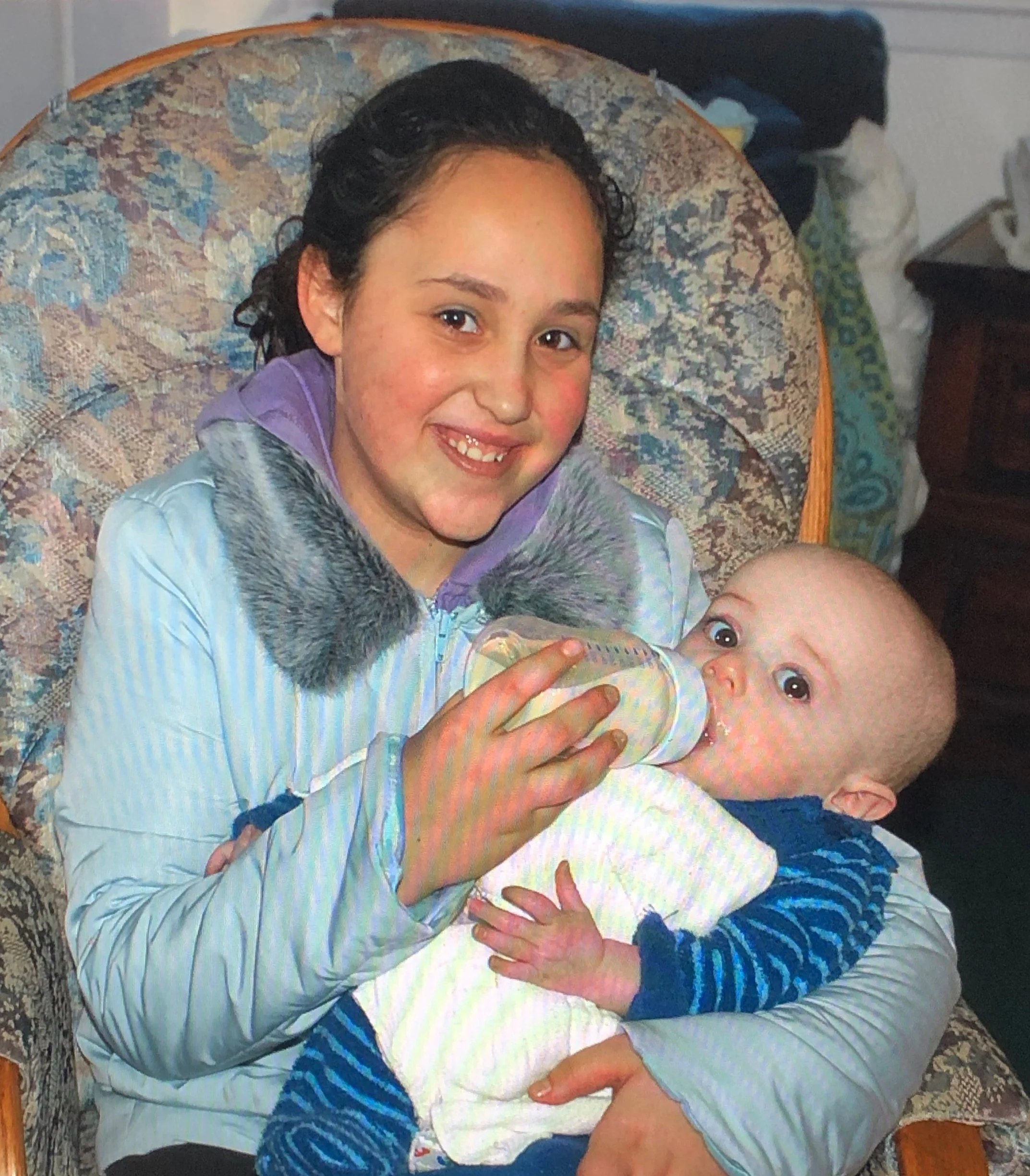A young girl with dark curly hair smiling, bottle-feeding a baby boy with light hair, sitting on a patterned armchair in a cozy room.