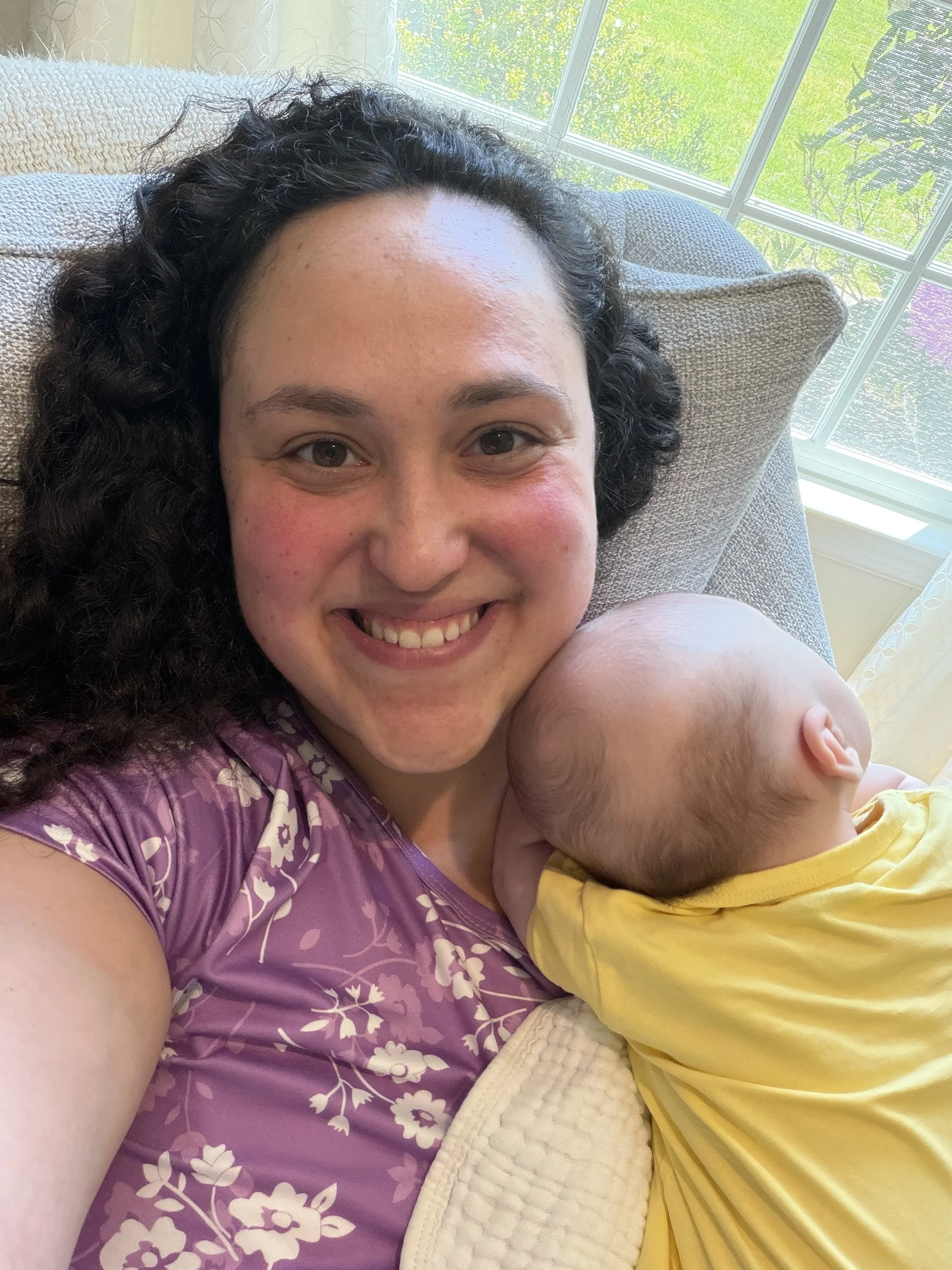 A woman with dark, curly hair smiling while holding a baby wearing a yellow outfit, sitting together on a couch near a window with green trees outside.