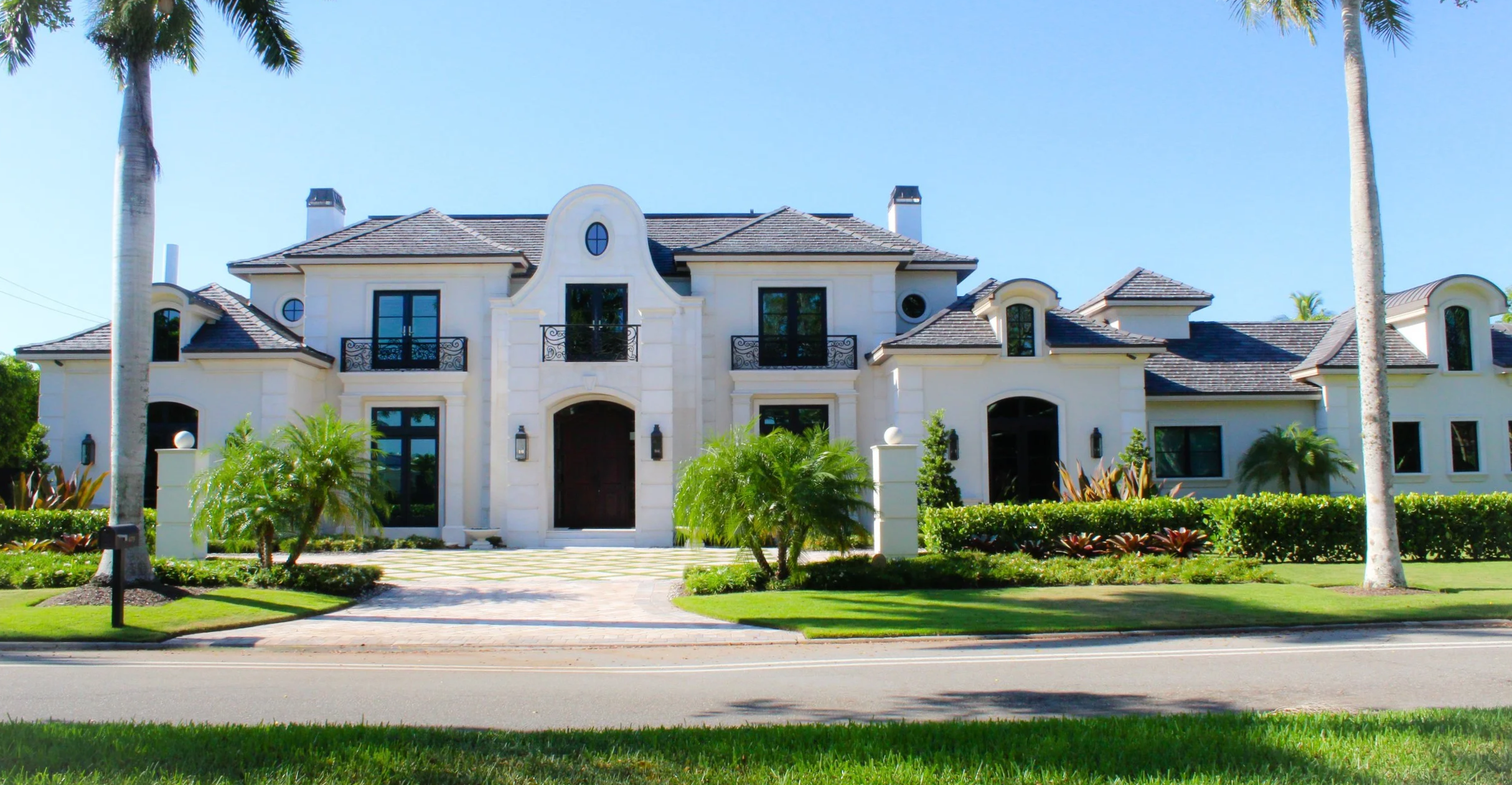 A large white mansion with dark brown doors, multiple windows, and balconies, surrounded by green shrubs, palm trees, and a well-manicured lawn, under a clear blue sky.