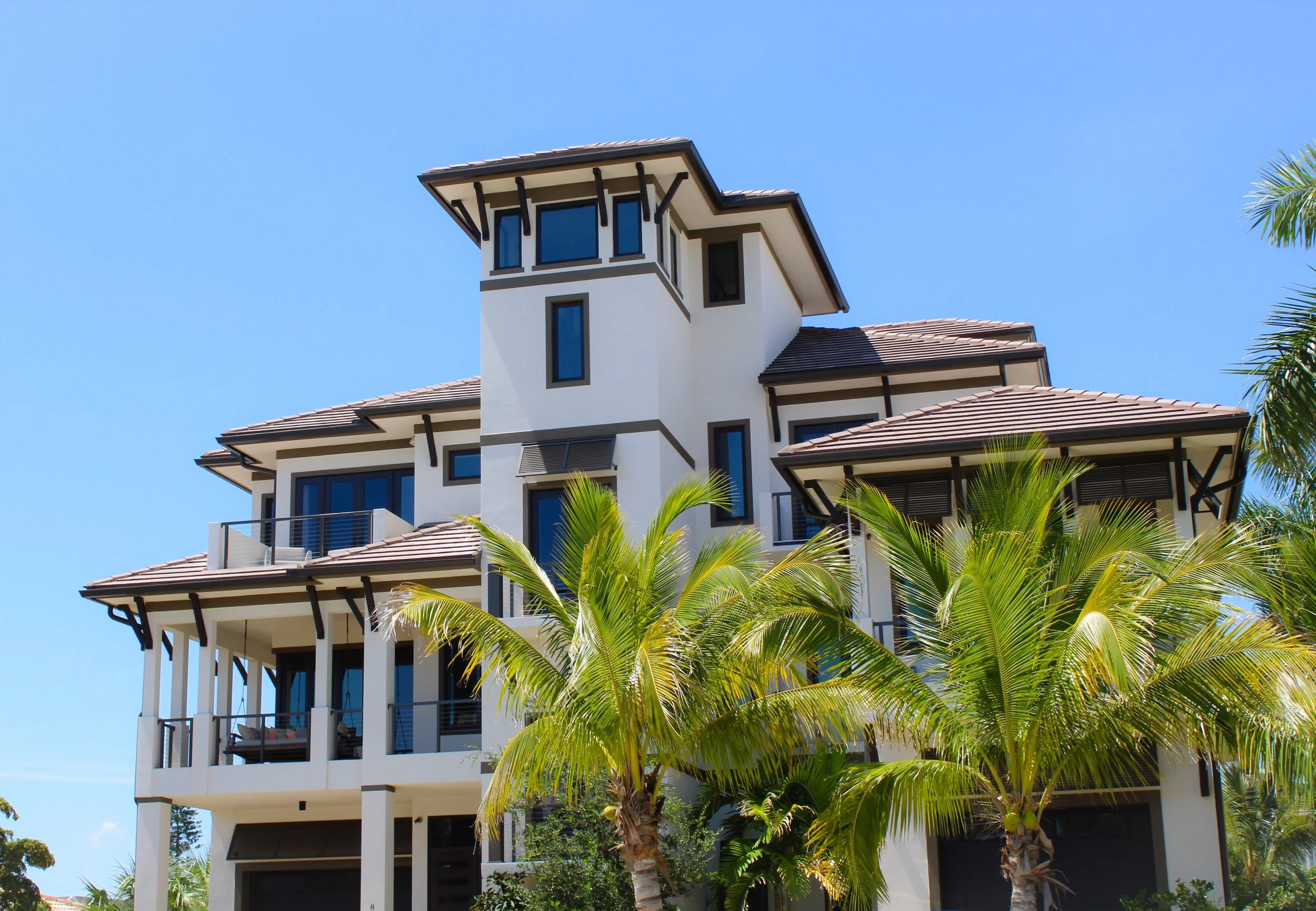 Modern multi-story house with balconies and a plethora of windows and doors, surrounded by palm trees under a clear blue sky.