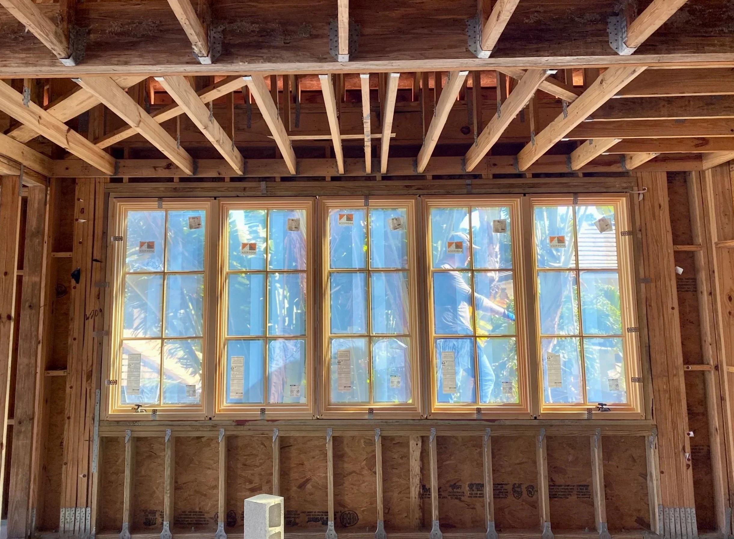 Interior view of a house under construction, showing wooden framing, a large multi-pane window, and two workers outside installing or inspecting the window.