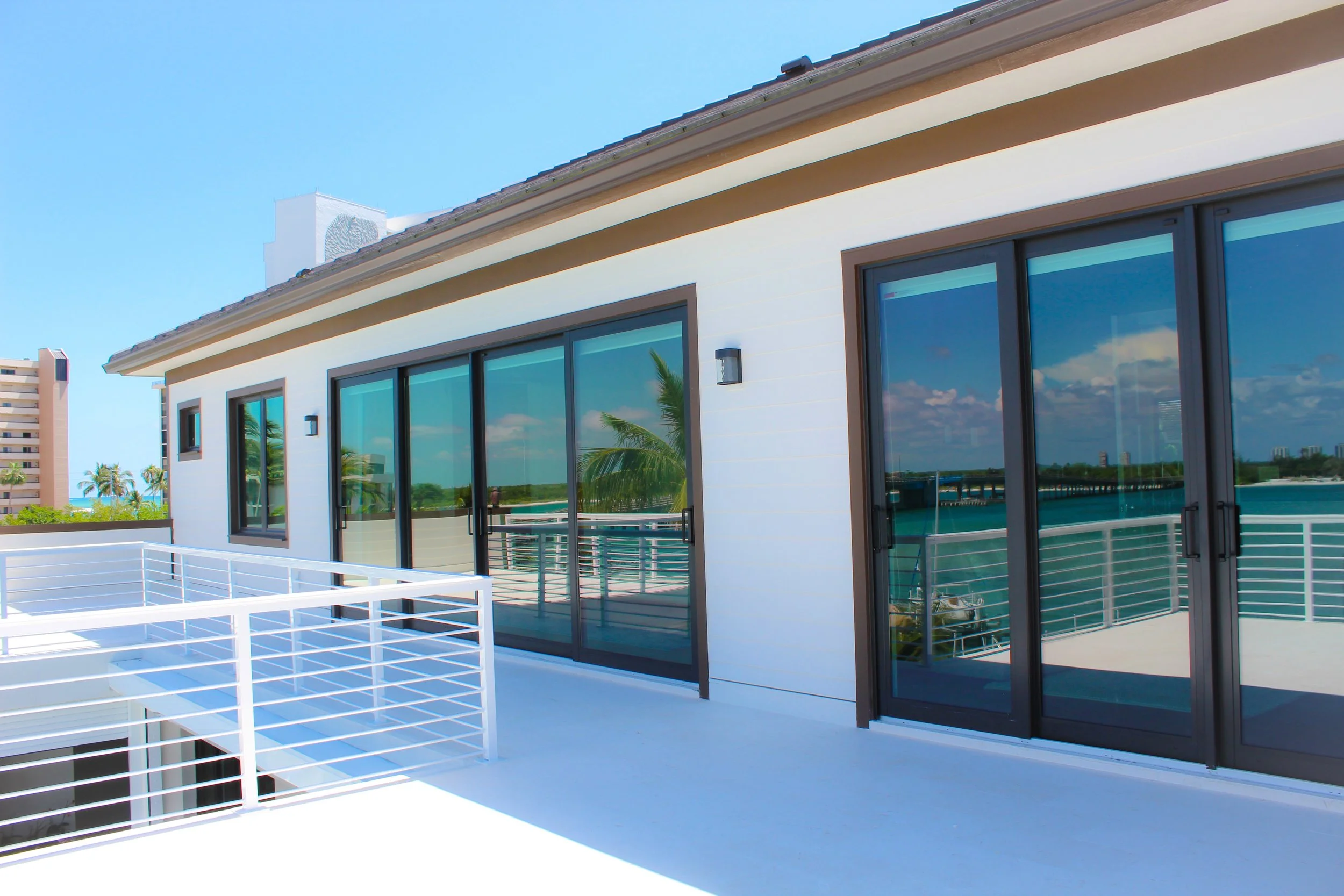 Balcony of a modern building with large glass sliding doors and a view of water and palm trees. Urban buildings are visible in the distance.