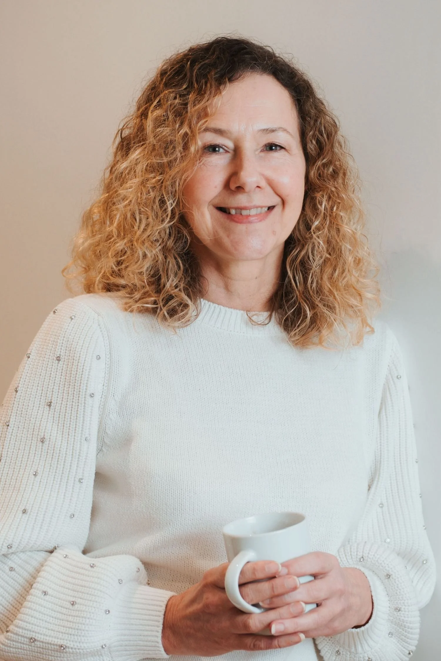 Woman with curly hair wearing a white sweater holding a white mug.