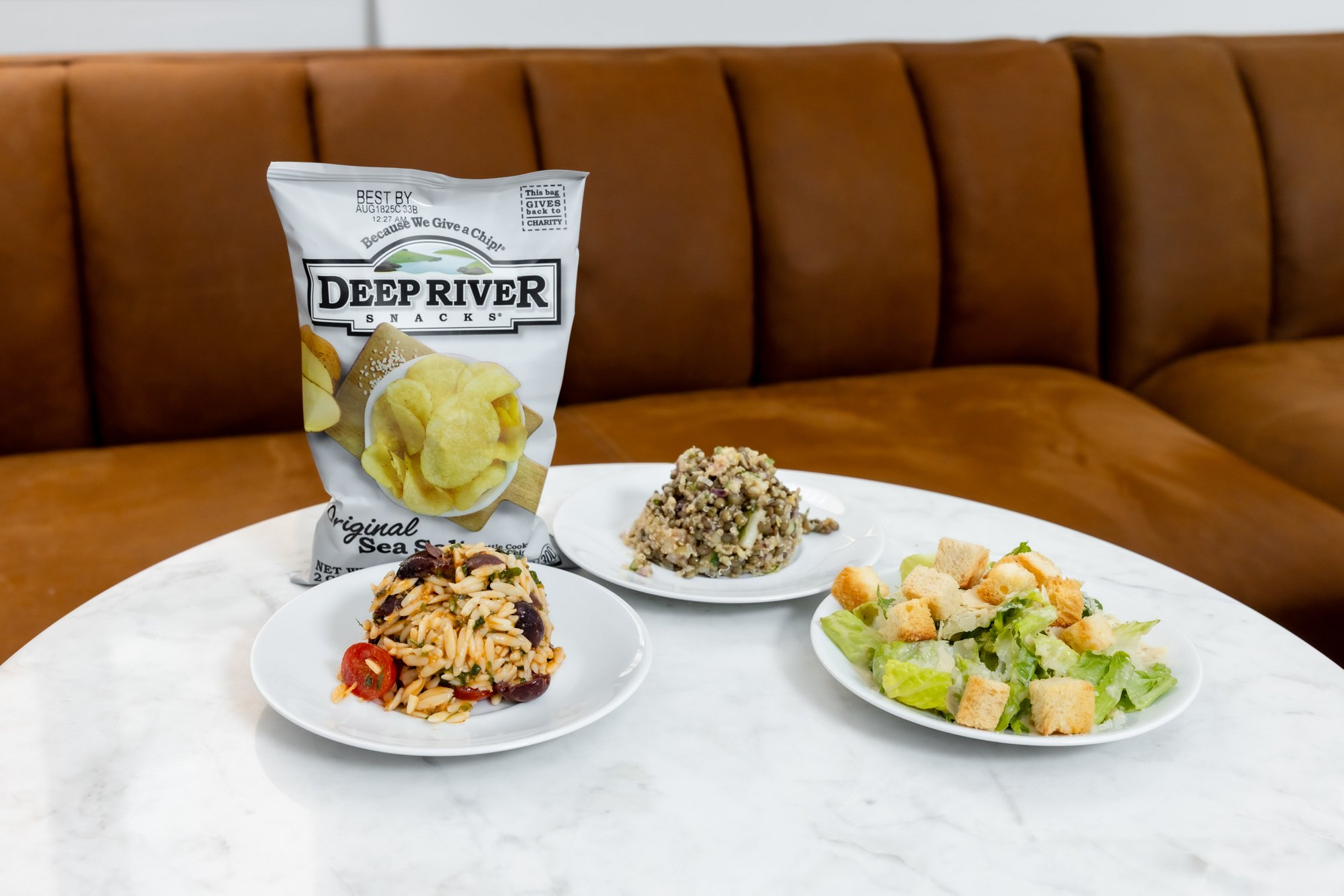 A marble table with a bag of Deep River Snacks potato chips, a pasta salad with cherry tomatoes, a grain salad with nuts, and a Caesar salad with croutons, in front of a brown leather sofa.