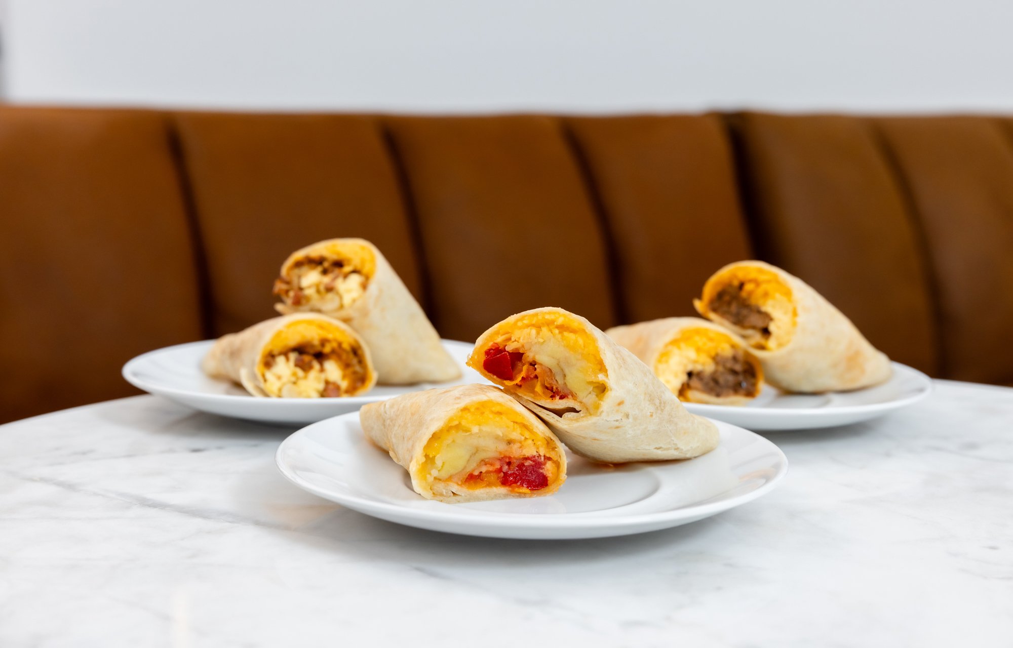 Plate of breakfast burritos with scrambled eggs, cheese, and sausage on a white marble table with a brown cushioned background.