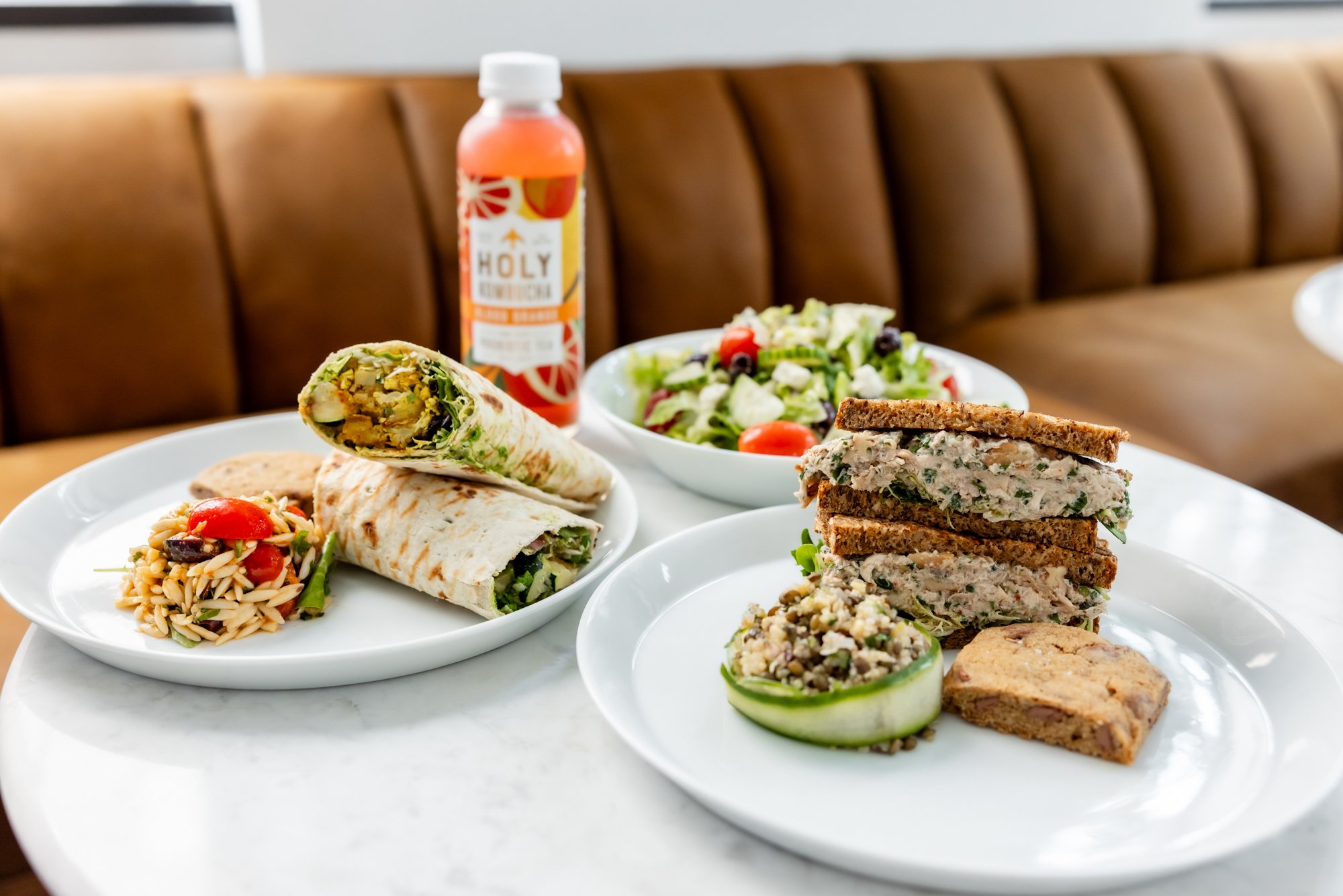 Plate with wraps, salad, couscous, and a bottle of orange juice on a marble table in a restaurant. A stacked sandwich with spread and greens, a cucumber roll with grains, and pasta salad are visible.