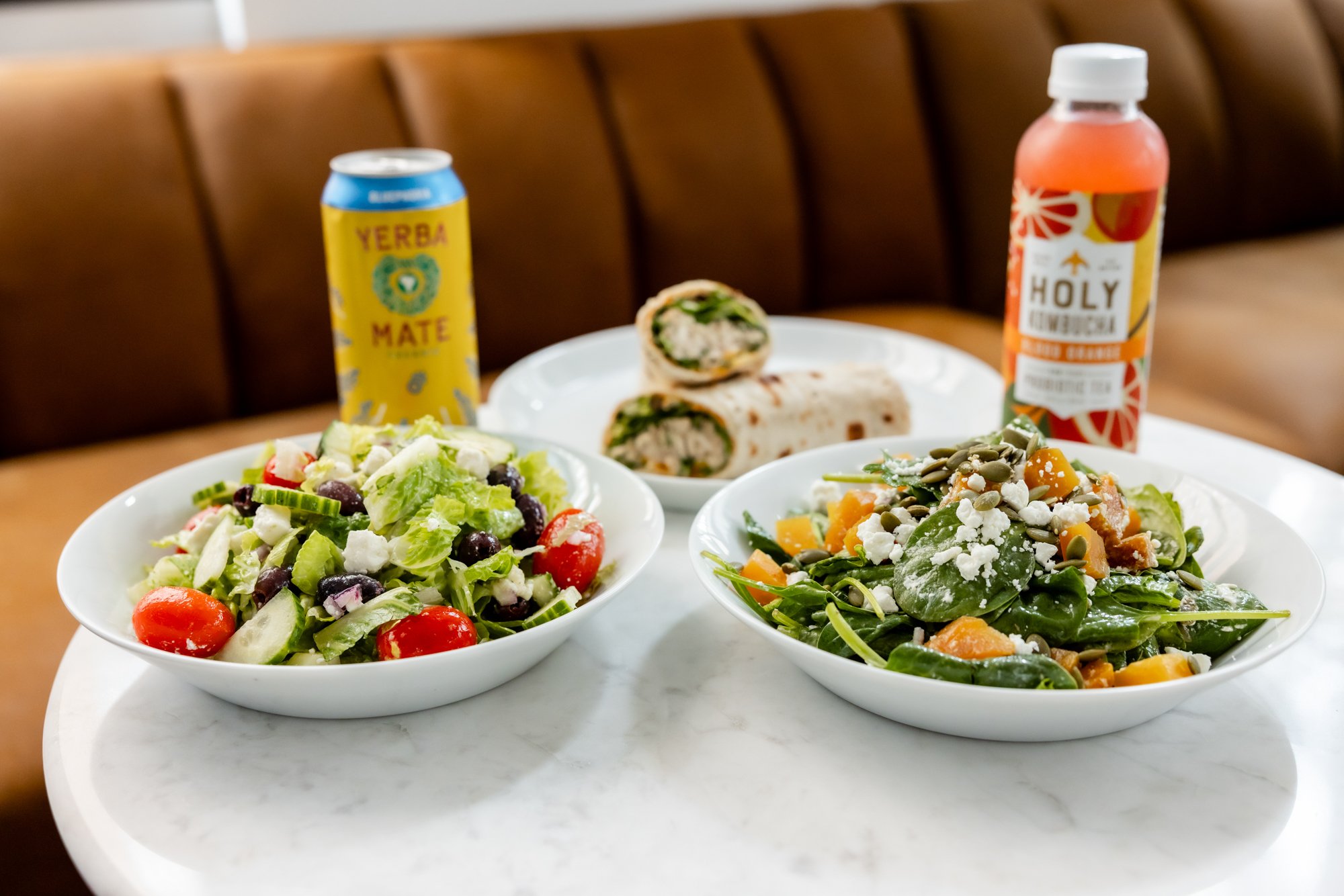 Salad, wrap sandwiches, canned drink, and bottled juice on a white marble table.