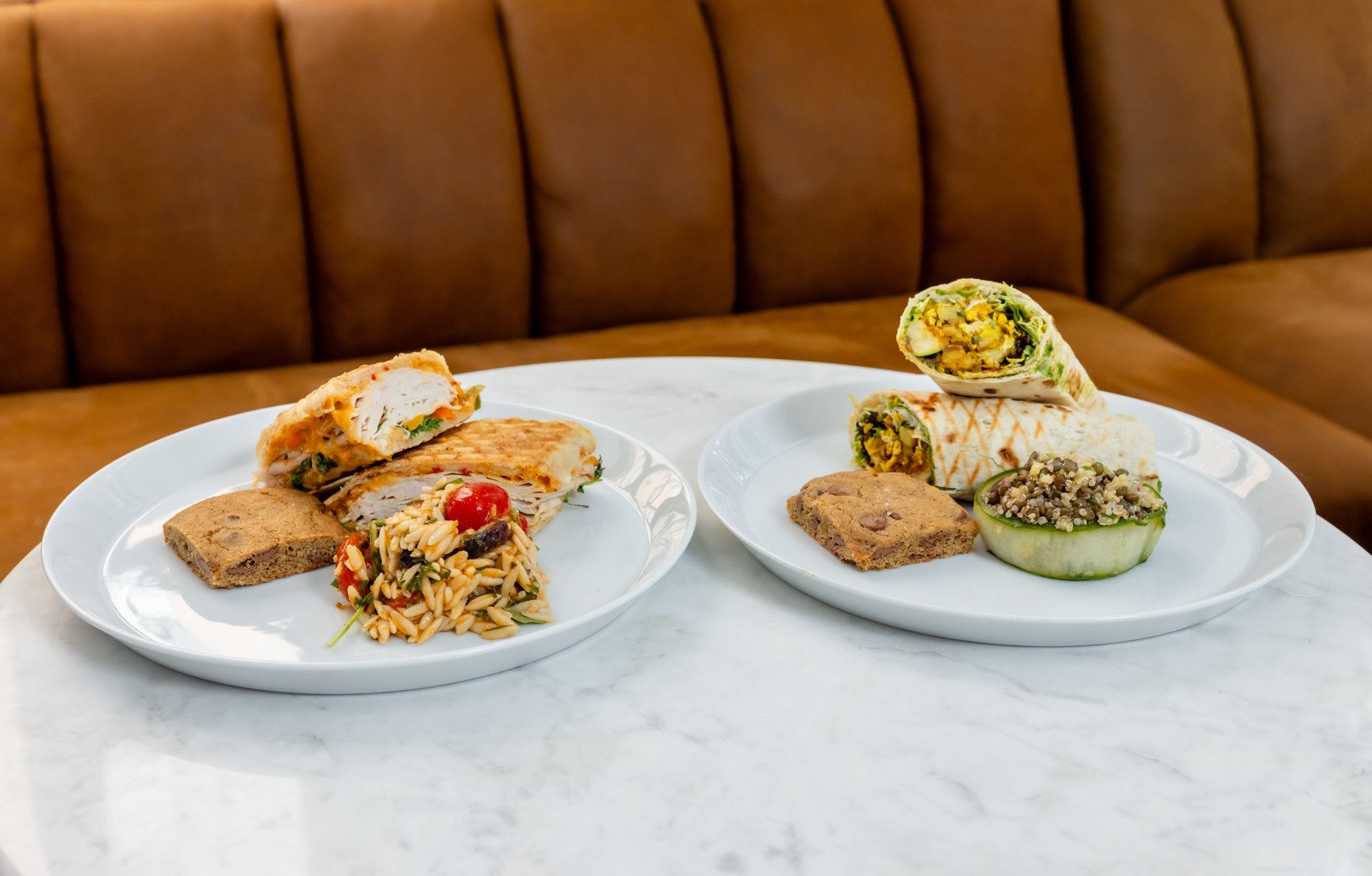 Two white plates with various wraps, salads, and cookies on a marble table in front of a brown cushioned bench.